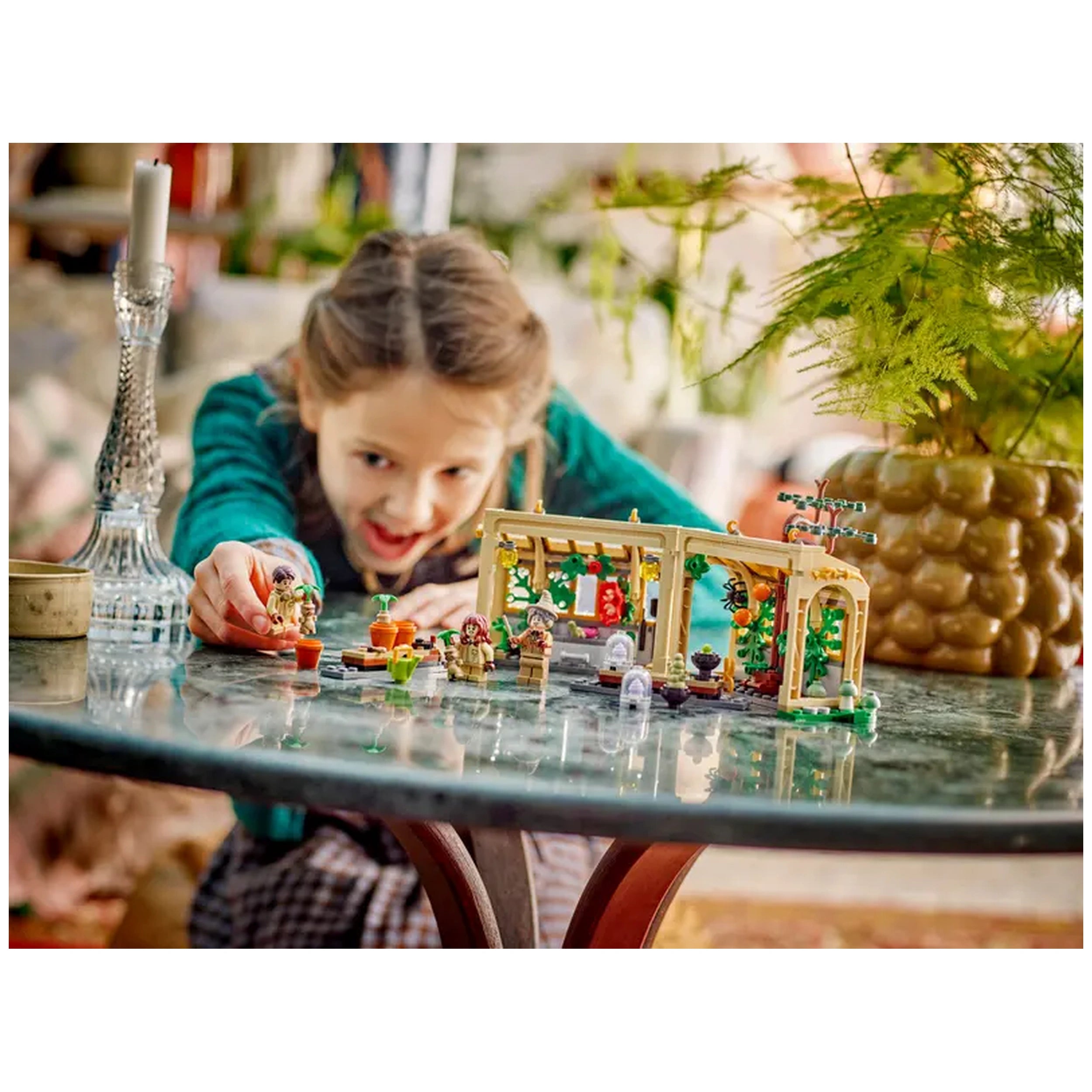 A child is excitedly playing with the LEGO Hogwarts™ Castle: Herbology Class set on a glass table, featuring detailed miniatures of plants and figures in a vibrant greenhouse.