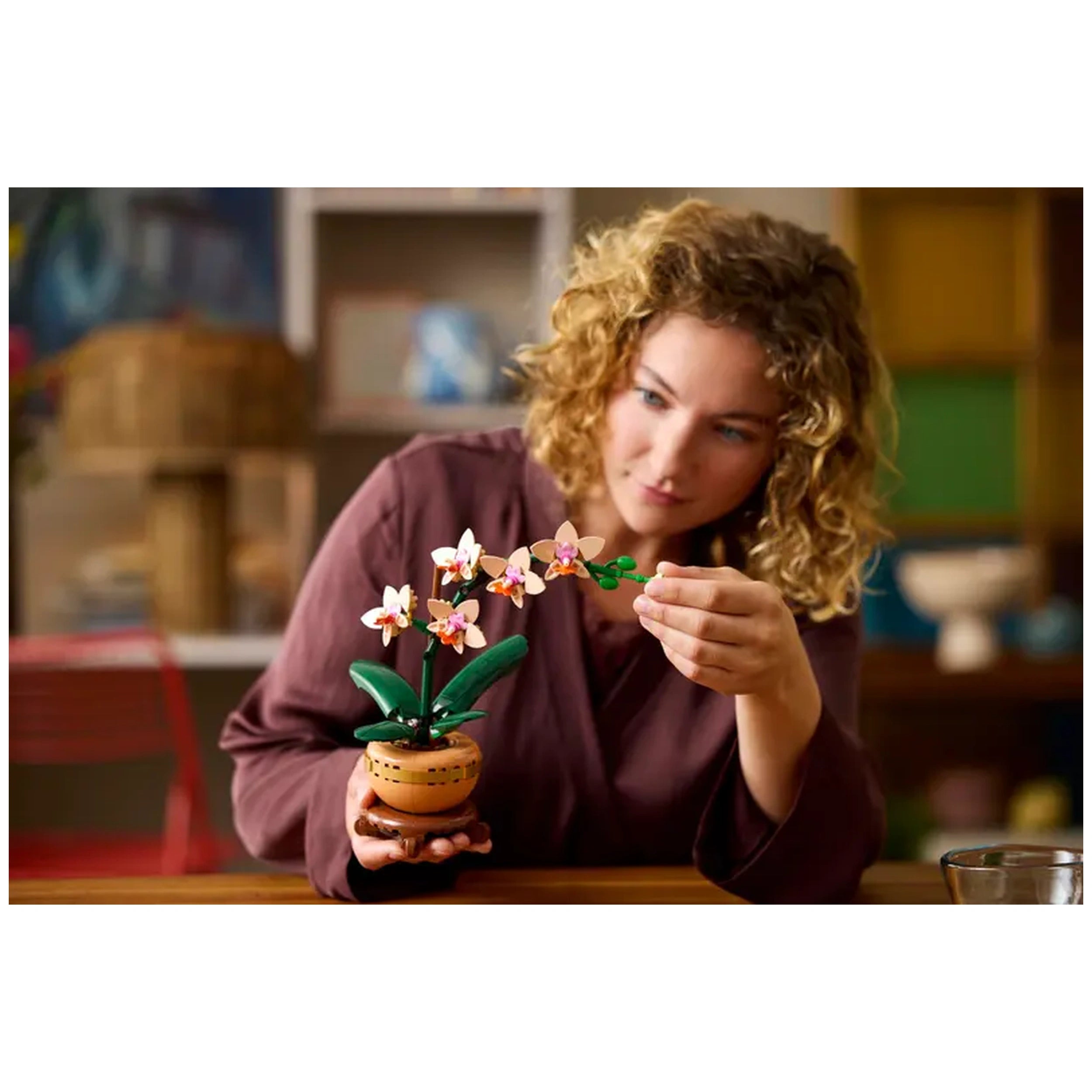 A woman with curly hair carefully adjusts a pink LEGO® Mini Orchid in a terracotta pot, showcasing its vibrant blooms and green leaves on a wooden table.