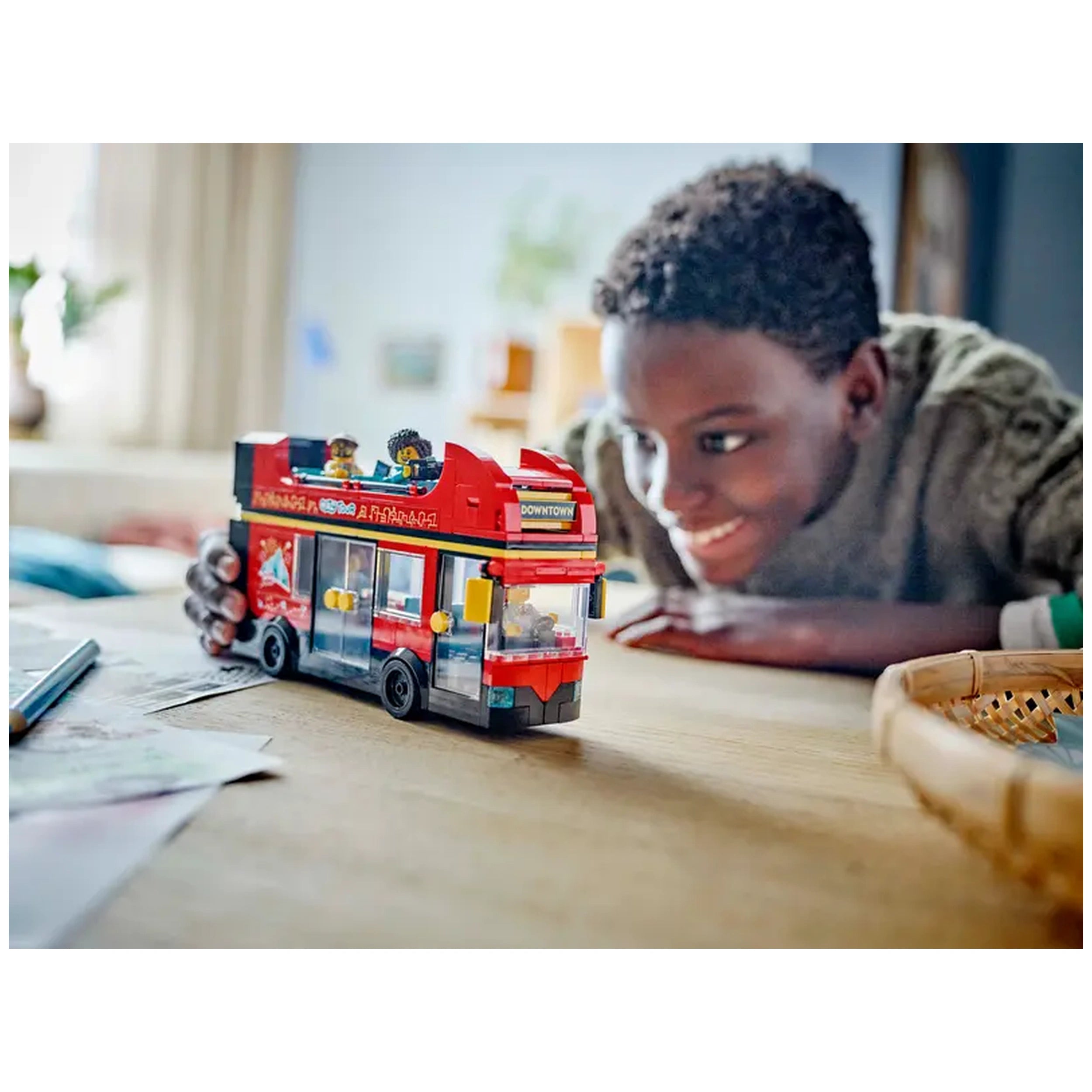 A child leans forward excitedly, examining a red LEGO® City Double-Decker Sightseeing Bus on a wooden table. The bus features a "Downtown" destination display and has two minifigures visible on the upper deck. Bright colors and playful details create an inviting scene for imaginative play.