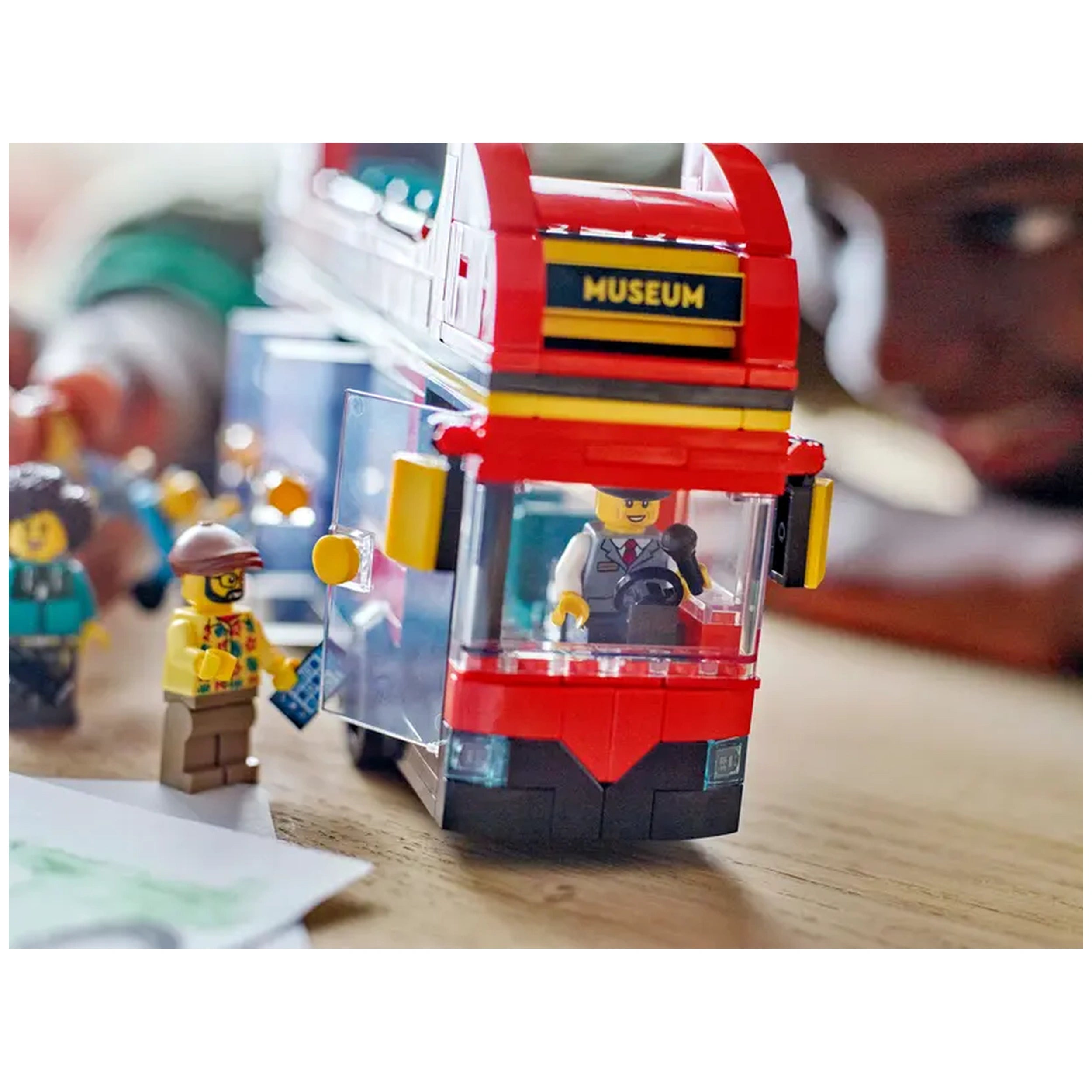 LEGO® City Double-Decker Sightseeing Bus in vibrant red, with a driver minifigure inside. A passenger in a floral shirt waits at the bus door. Various minifigures surround the scene, depicting a lively city atmosphere.