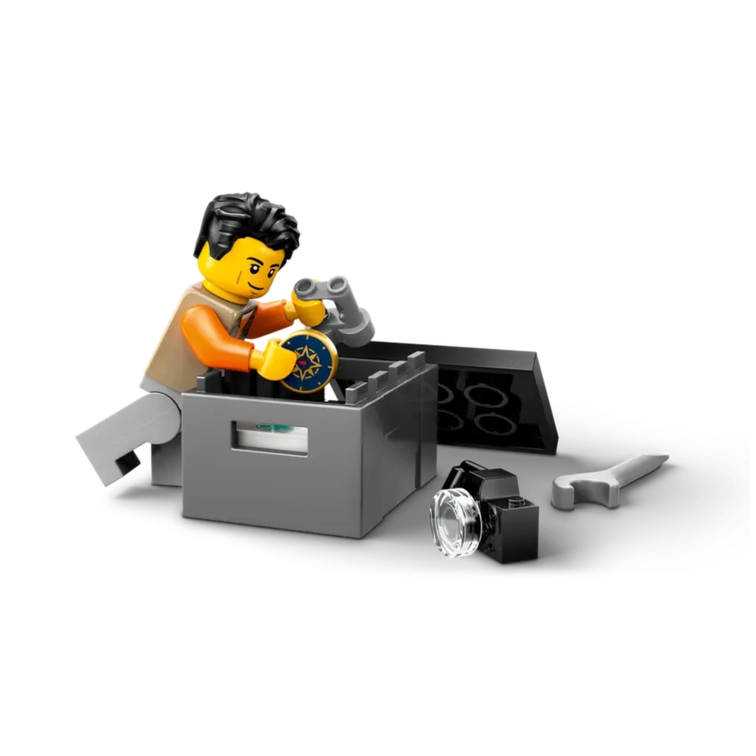 A LEGO figure in an orange shirt kneels beside a gray storage box, holding a compass and looking at an off-road truck part. Tools and accessories are scattered nearby.