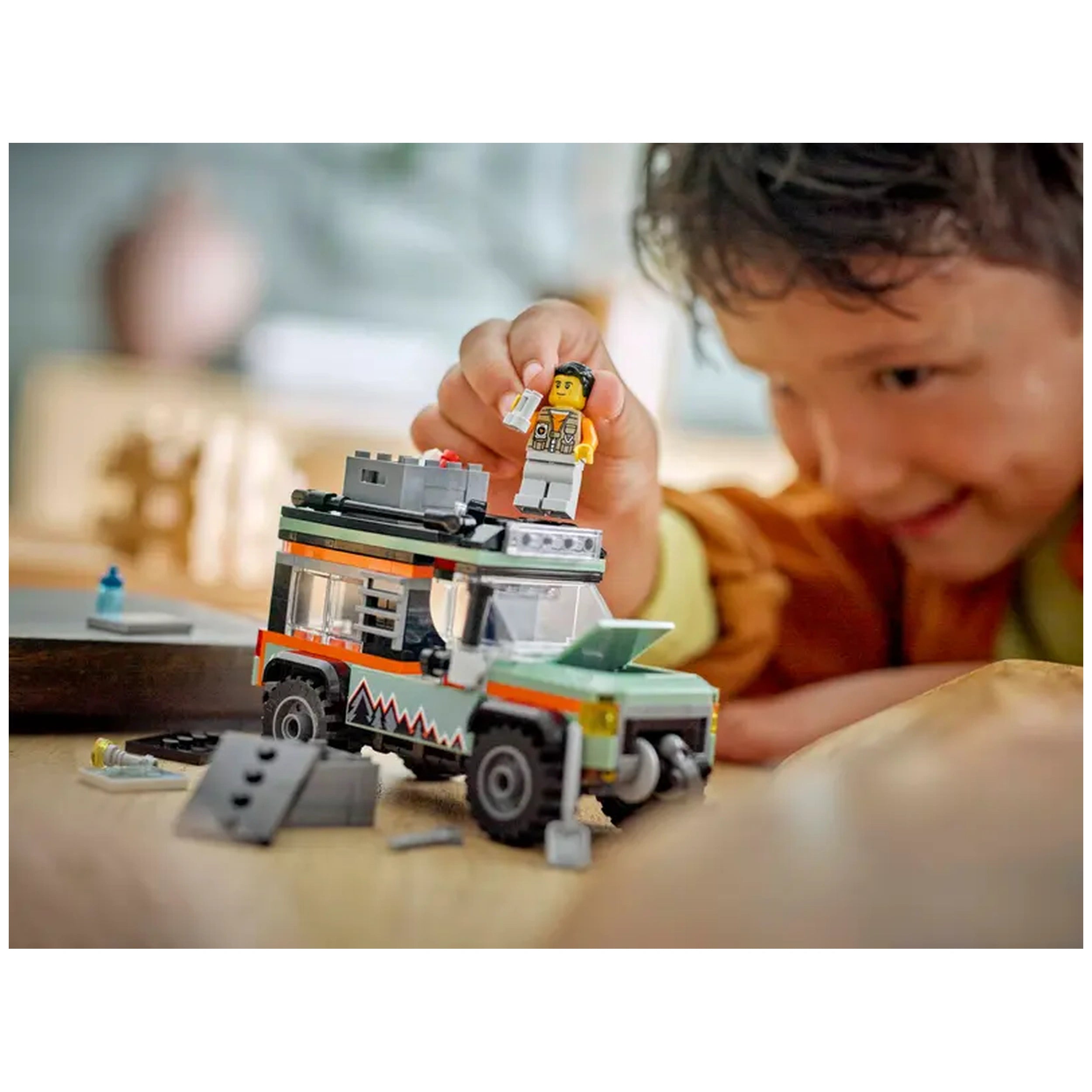 Young child playing with the LEGO® City Off-Road Mountain Truck, holding a mini figure while exploring accessories scattered on a wooden table.