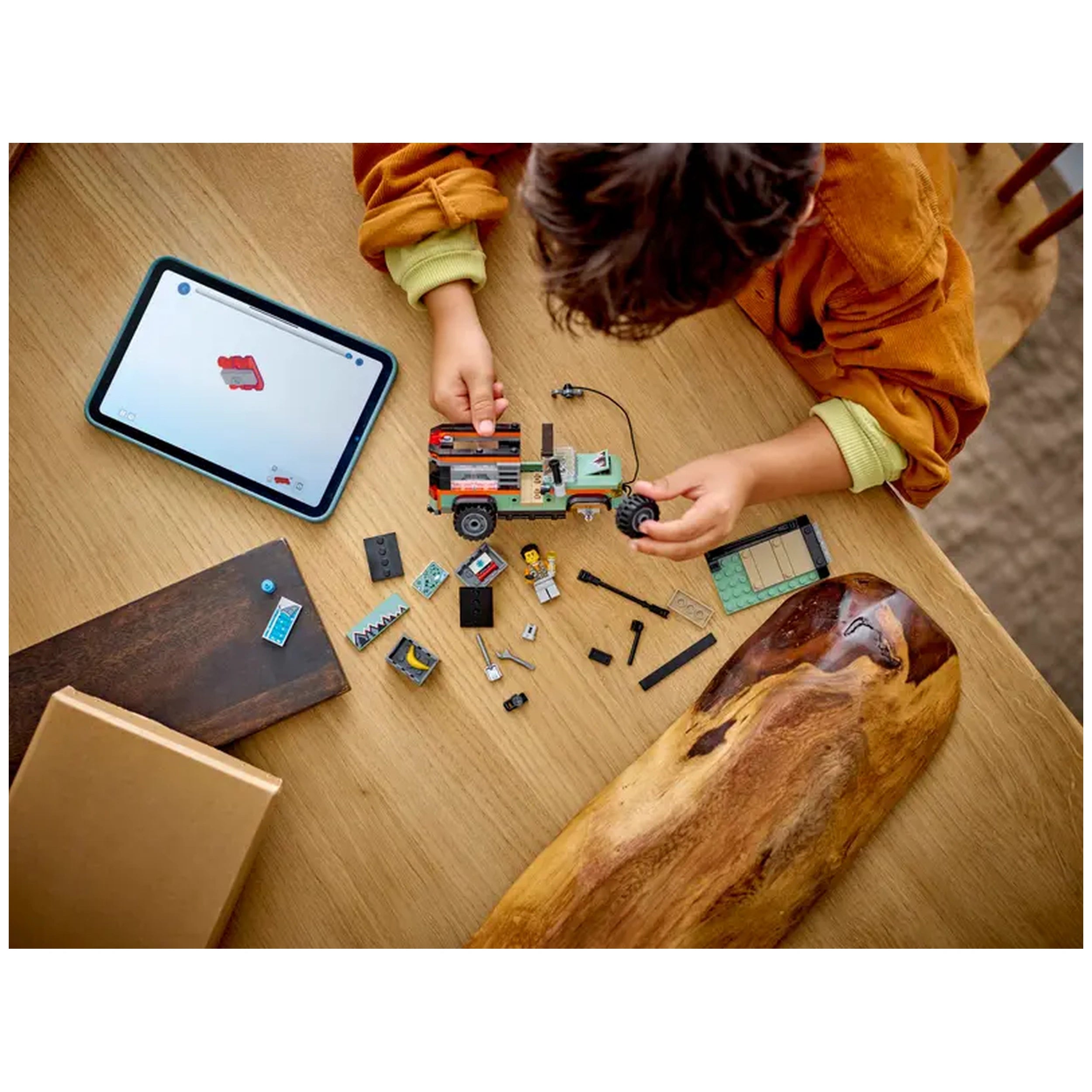 Child assembling the LEGO® City Off-Road Mountain Truck on a wooden table, surrounded by colorful building pieces and a tablet displaying instructions.