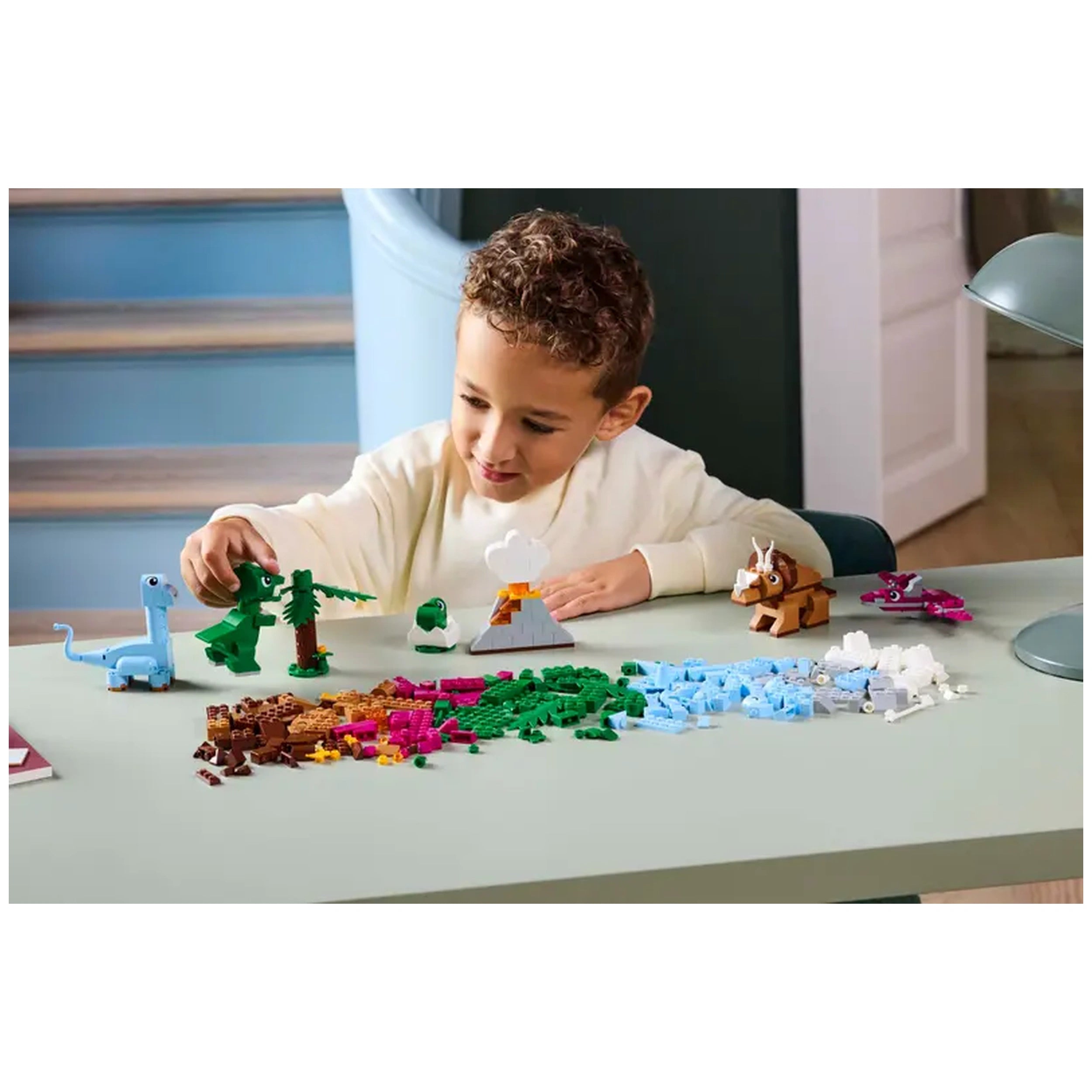A young boy in a cream sweater joyfully builds with colorful LEGO bricks on a light green table, featuring dinosaur figures, a volcano, and lush greenery.