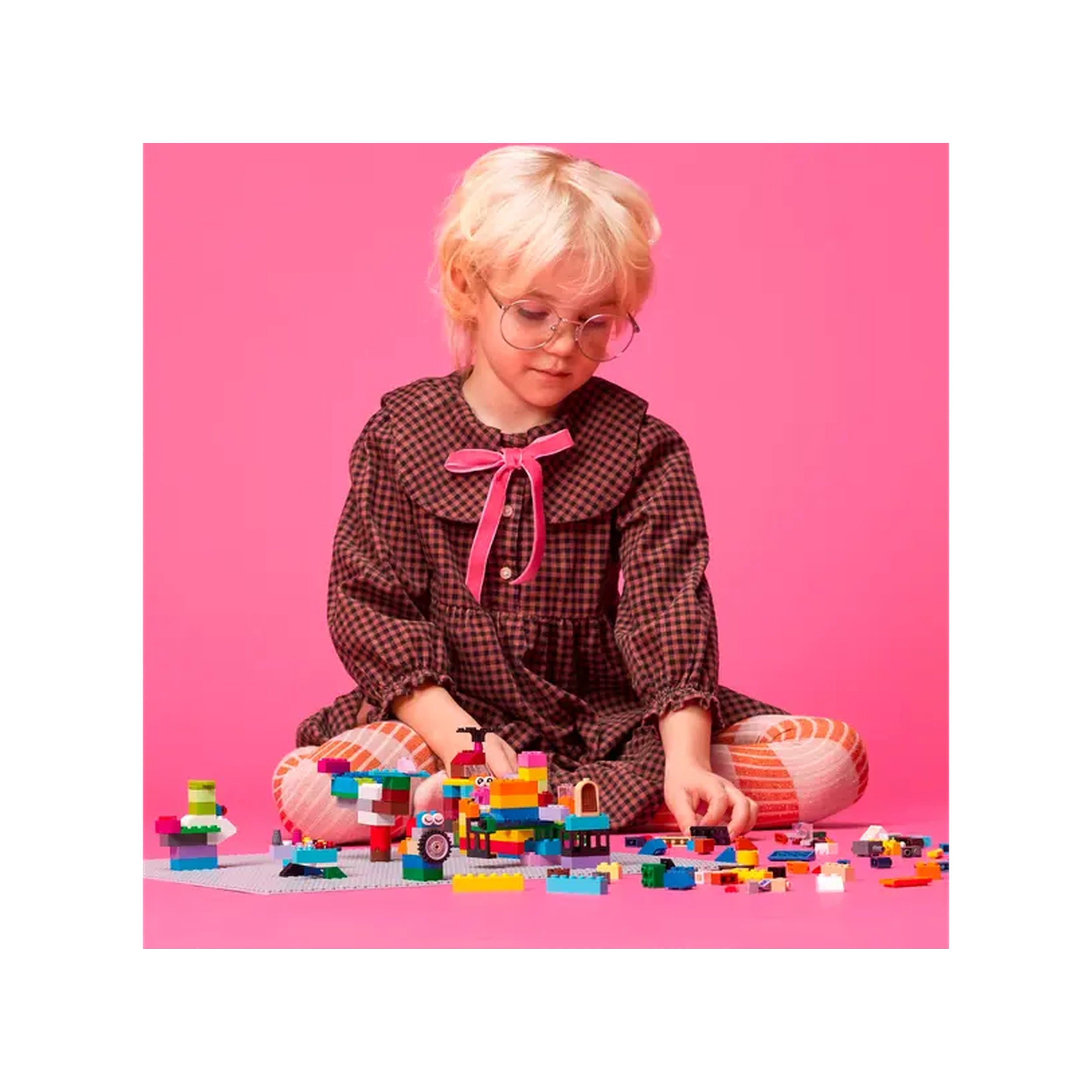 A child with short blond hair, wearing a plaid dress with a pink bow, sits on a pink background, focused on building with colorful LEGO bricks scattered around a gray baseplate.