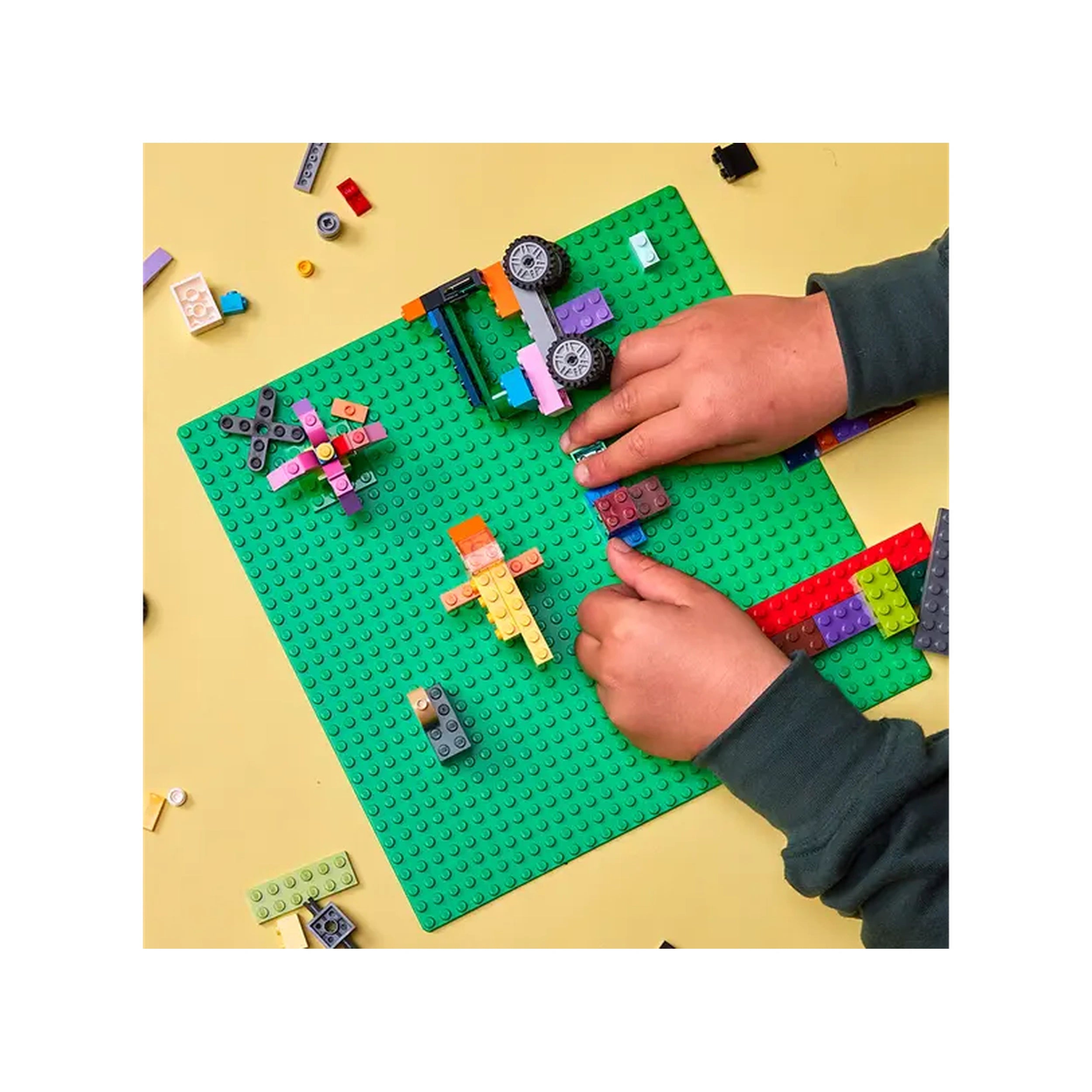 A child's hands build on a vibrant green LEGO baseplate, surrounded by colorful bricks and small creations, fostering creativity.