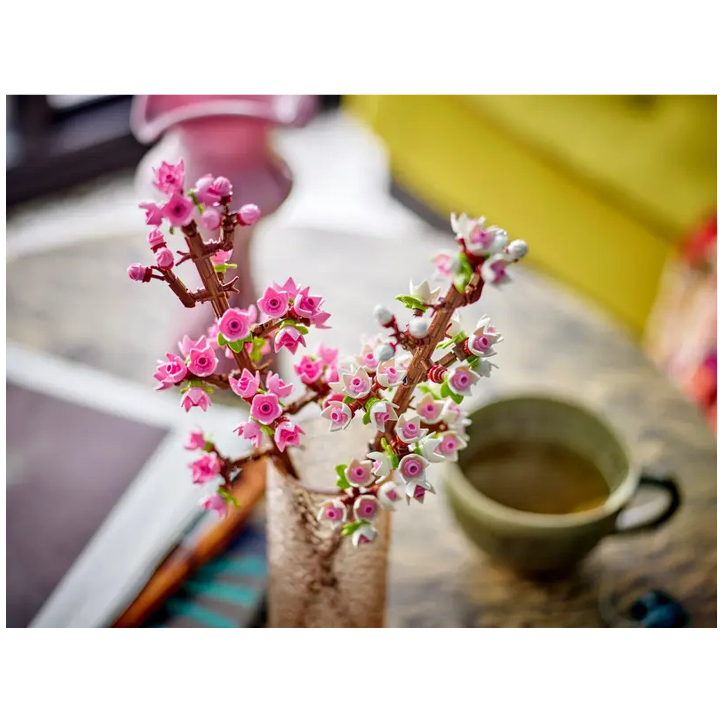 Delicate LEGO® Creator Cherry Blossom twigs in pink and white arranged in a clear vase, with a green cup nearby, set on a textured table.