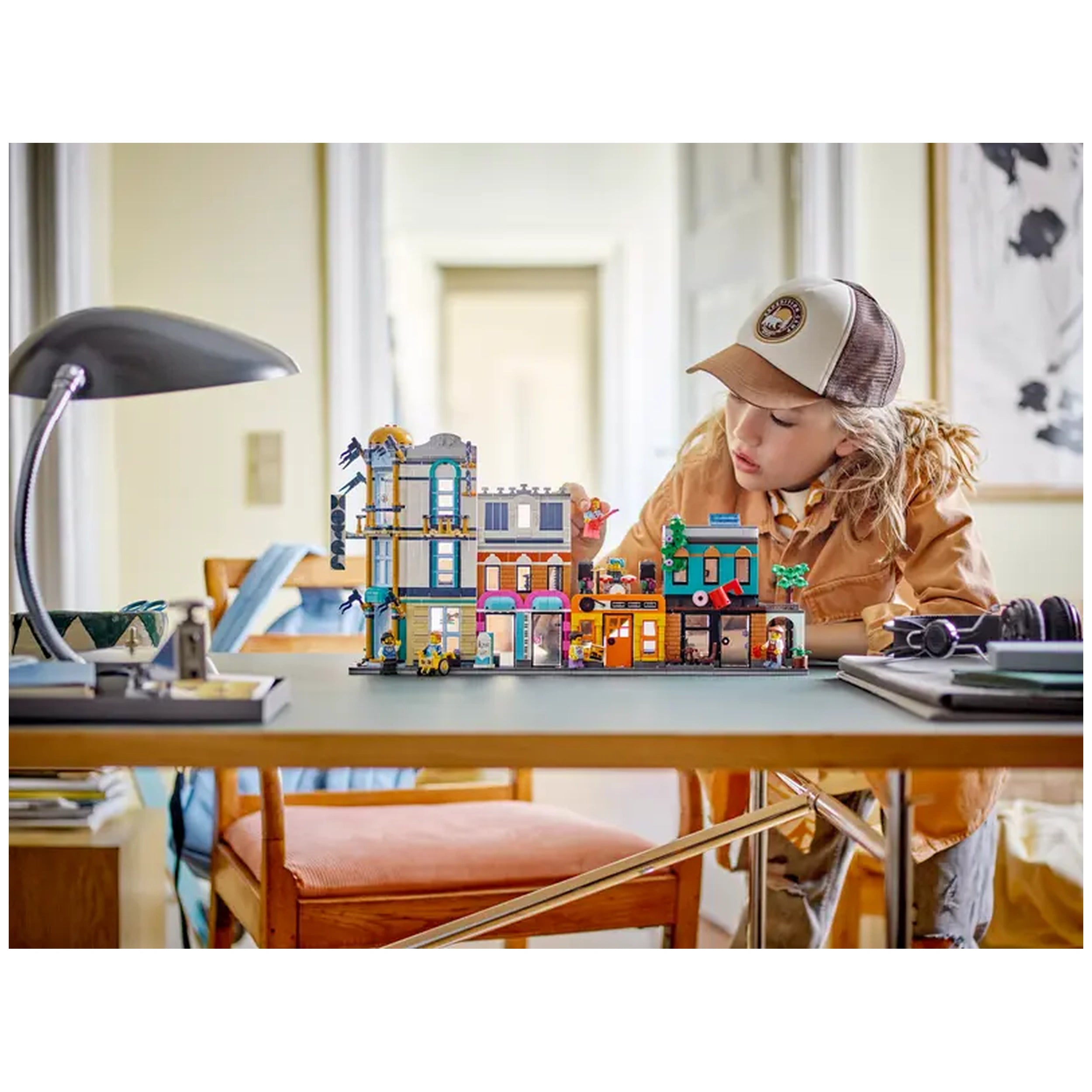 A focused child studies the LEGO Creator 3in1 Main Street set on a table, showcasing a vibrant cityscape with a hotel, music store, café, and record store. The child, wearing a cap and an orange jacket, engages creatively with the colorful buildings amidst a cozy indoor setting.