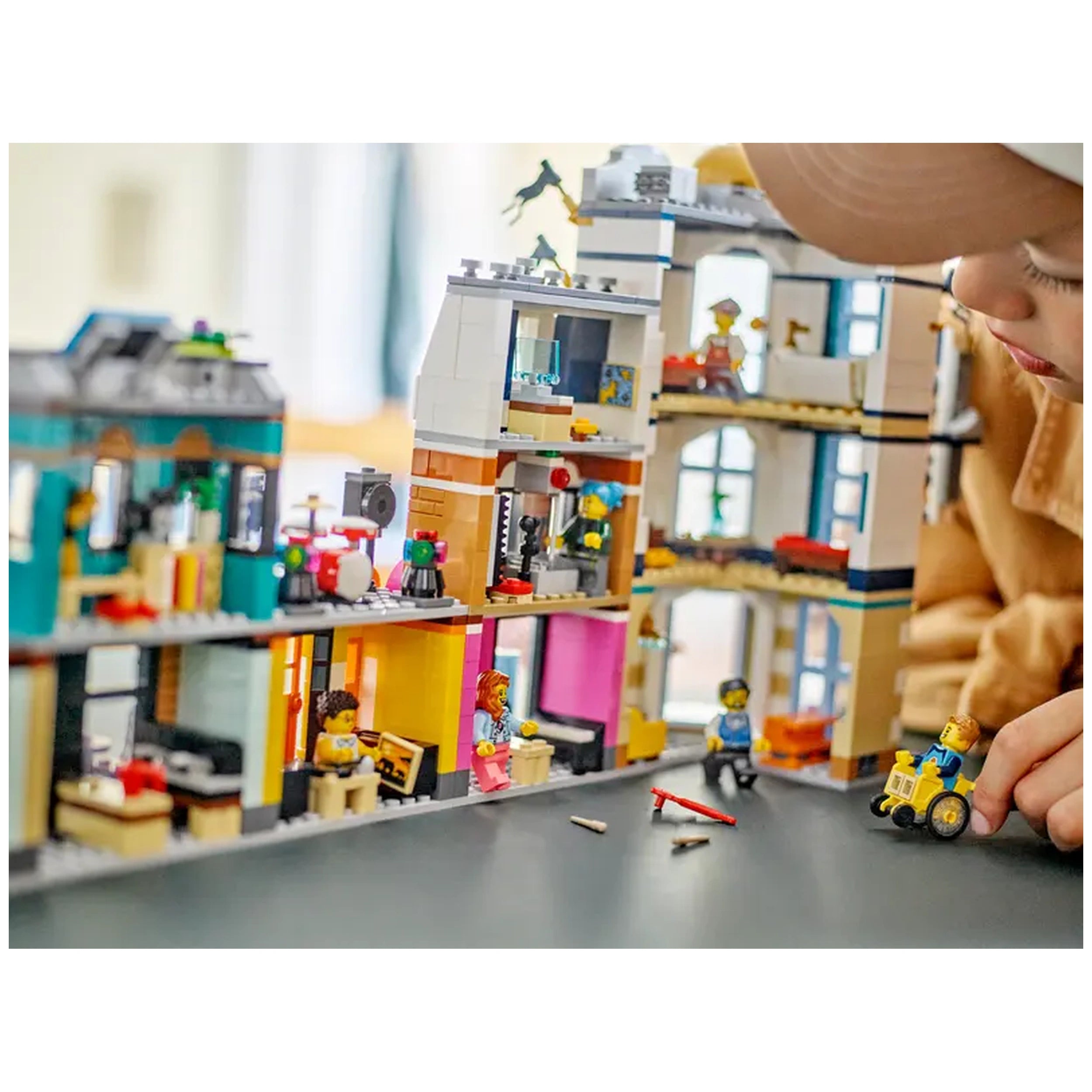 Colorful LEGO® Creator 3in1 Main Street set with detailed buildings, figures, and a child assembling a scene on a table.