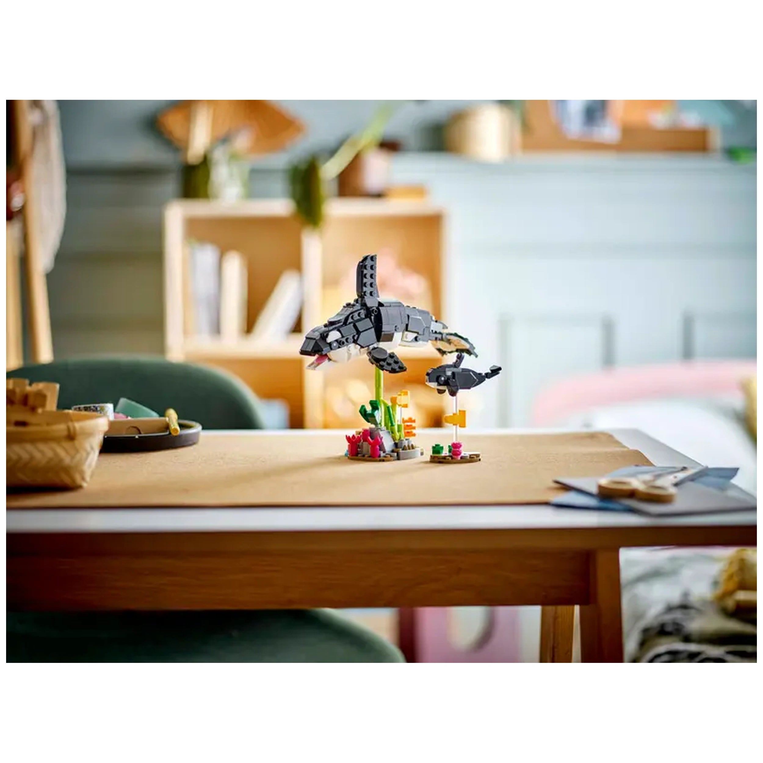 A LEGO Creator set displaying a black and white orca and its calf soaring above a colorful underwater scene with plants and coral on a table, set in a cozy room.