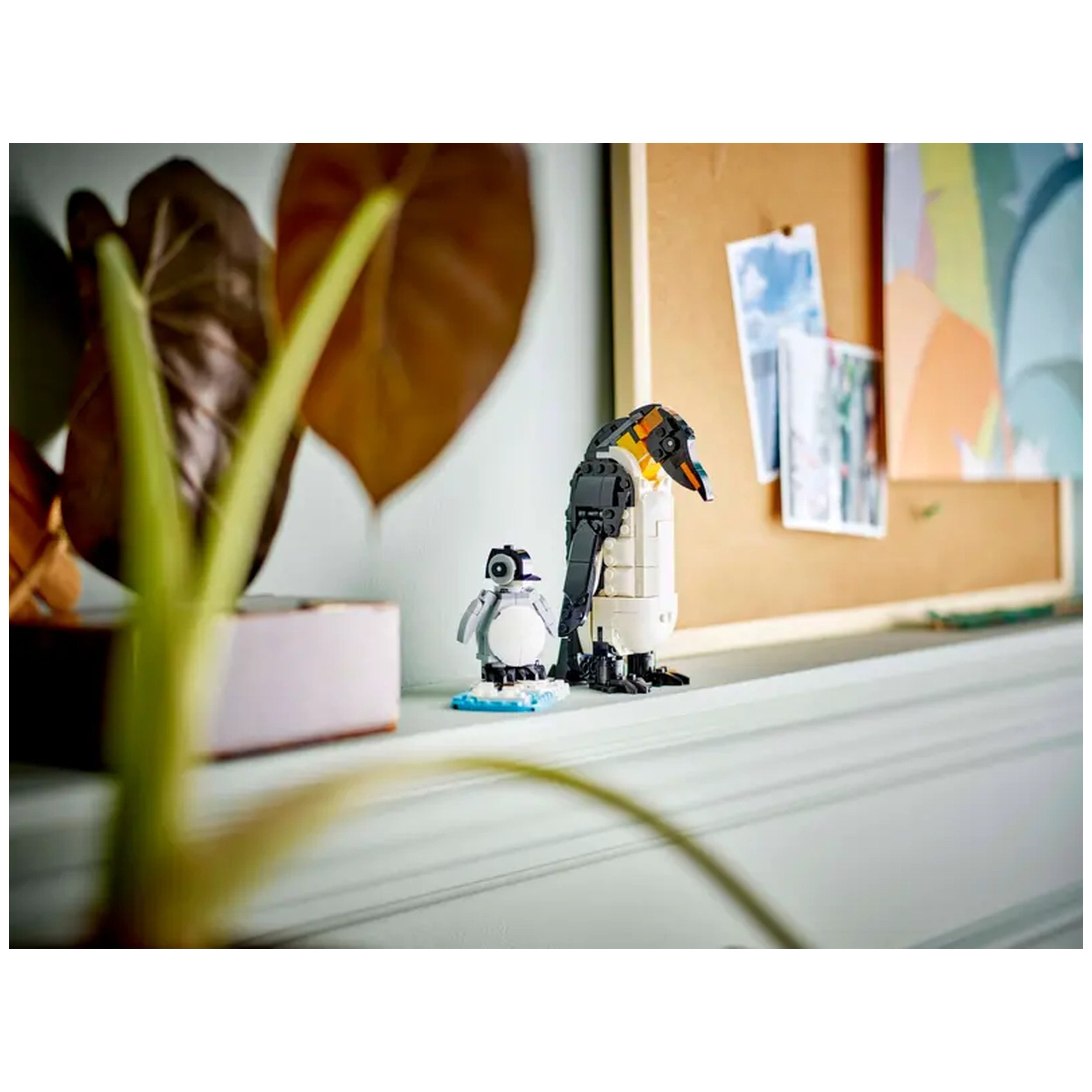 A black and white LEGO penguin stands on a blue ice base next to a larger LEGO emperor penguin with orange accents. Green leaves partially frame the scene, with a corkboard displaying various images in the background.