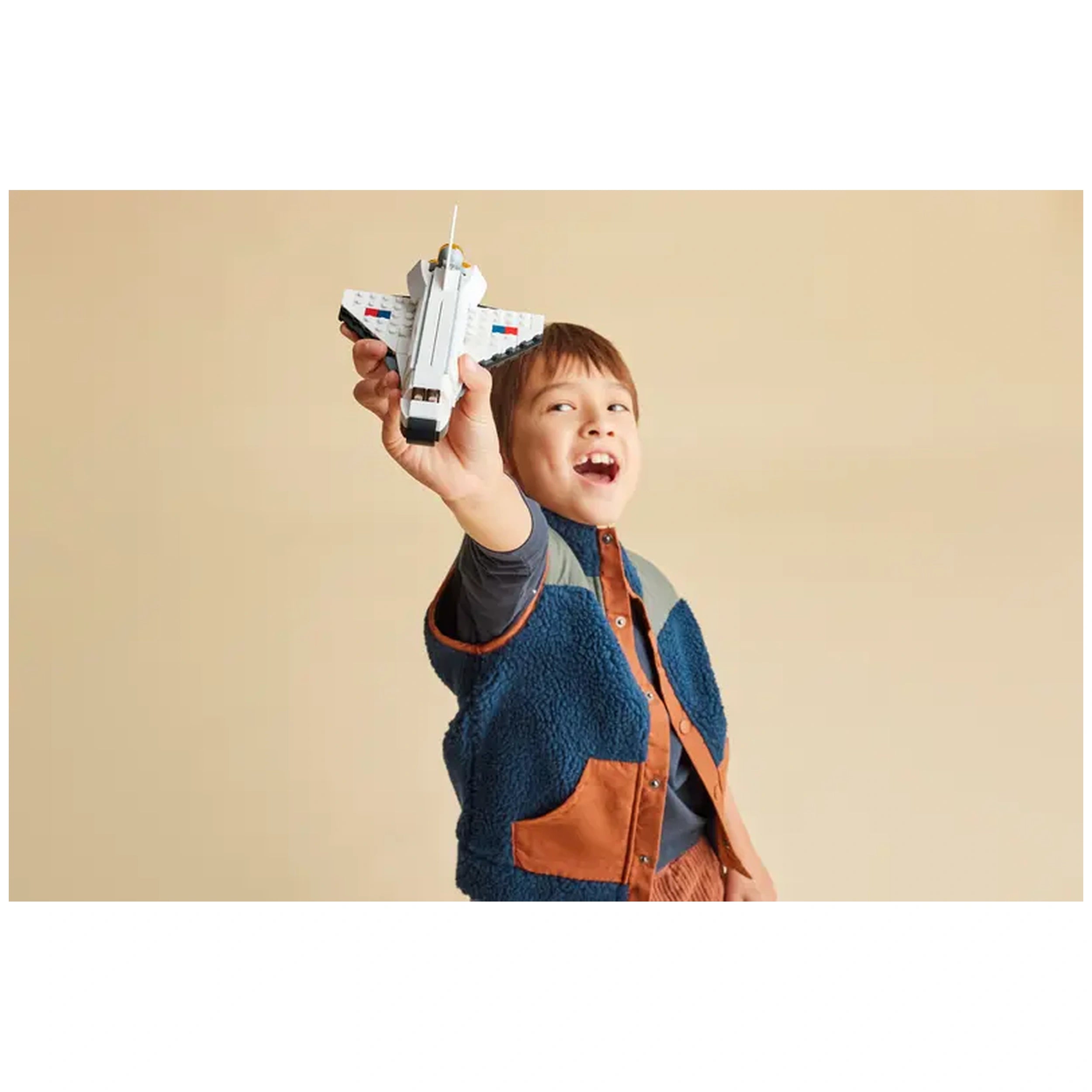 A young boy excitedly holds the LEGO Creator 3in1 Space Shuttle toy above his head, smiling joyfully. He wears a blue and brown fleece jacket. The background is a soft beige color, enhancing the playful atmosphere.