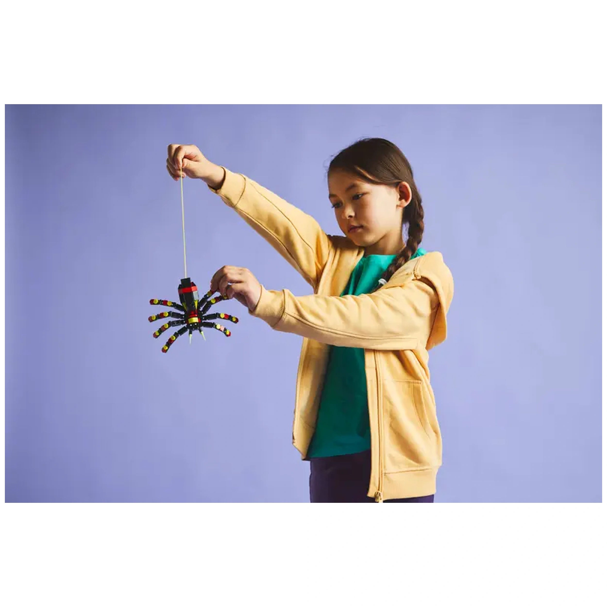 A child in a yellow hoodie holds a colorful LEGO Surprising Spider, showcasing its articulated legs and vibrant design against a soft purple background.