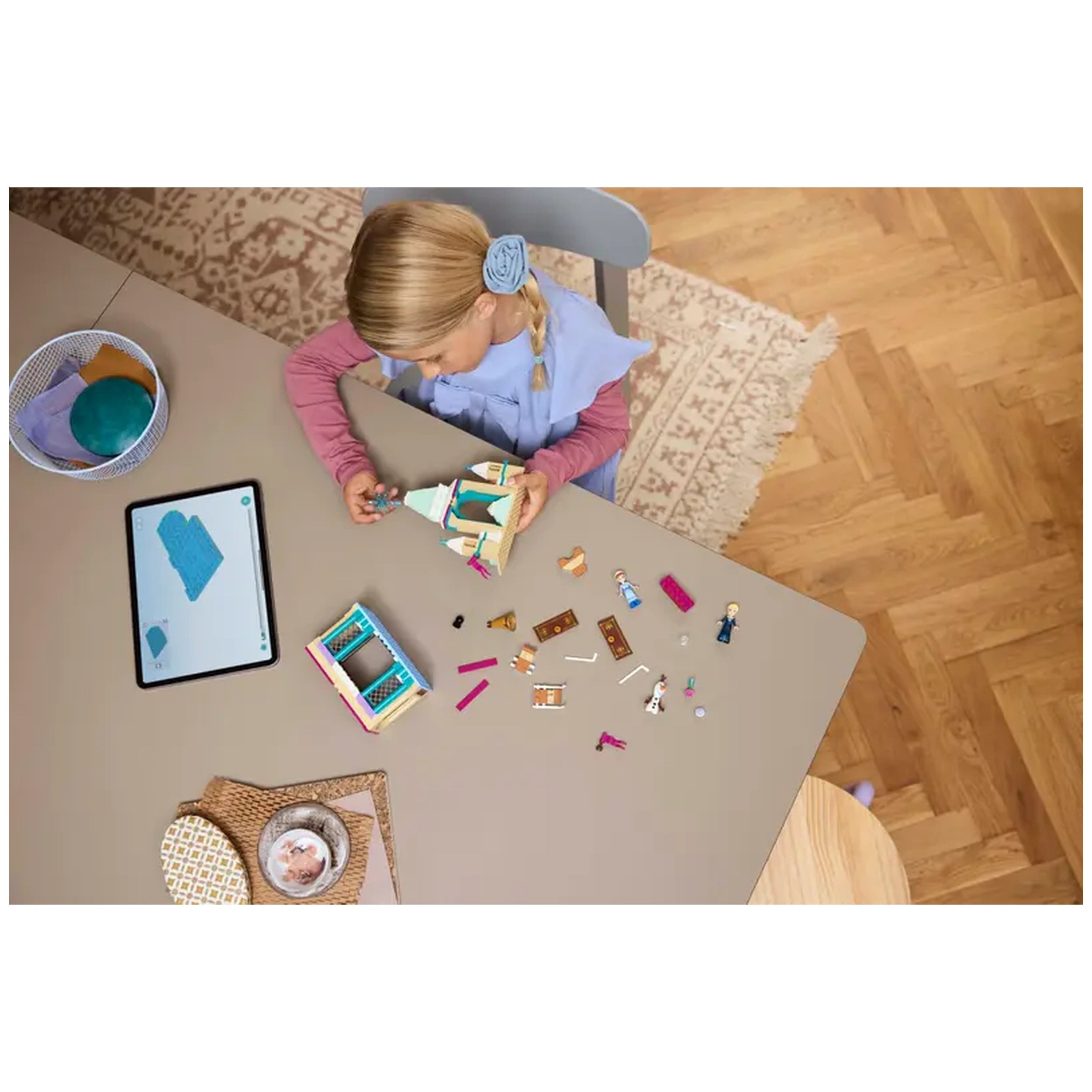 A child in a blue dress and pink sleeves focuses on building a LEGO Frozen castle, surrounded by colorful pieces and mini-doll figures on a beige table. An tablet displays instructions, with a cozy rug and wooden floor in the background.
