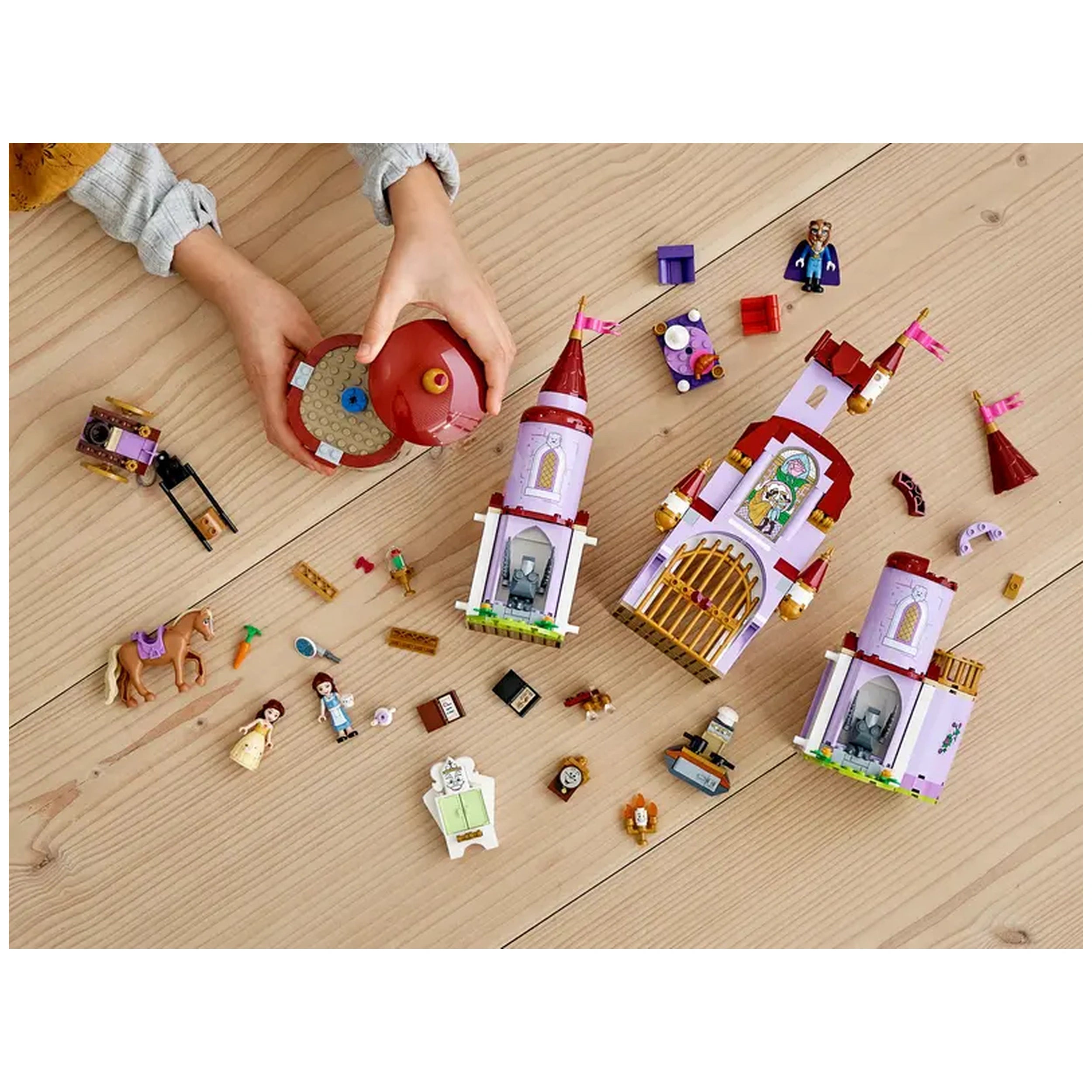 Colorful LEGO® Disney Belle and the Beast’s Castle construction set scattered on a wooden table, featuring castle pieces, characters, and accessories. A child's hand holds a red spinning top, while two versions of Belle and various accessories are visible nearby.