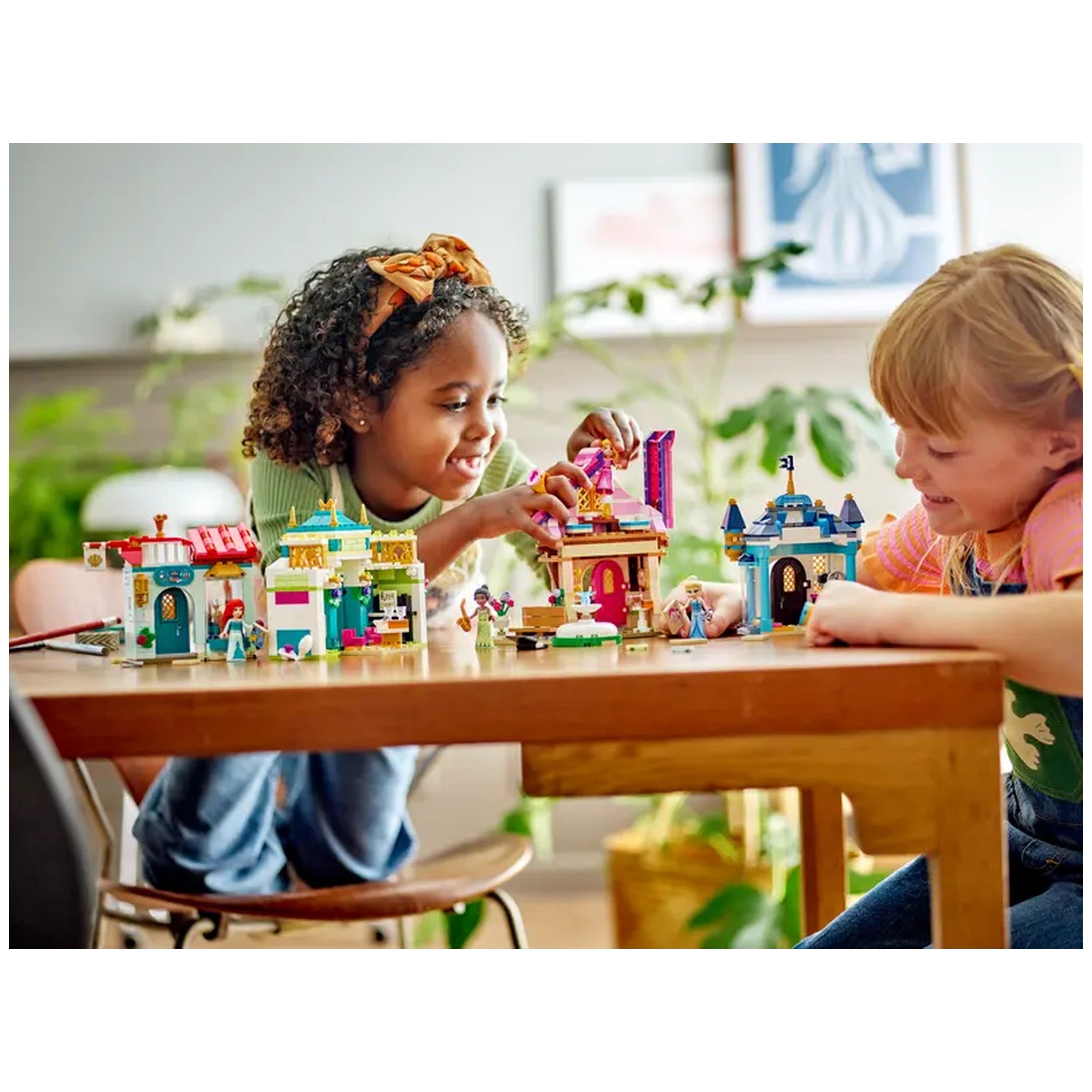 Two children joyfully playing with the LEGO Disney Princess Market Adventure set, featuring colorful toy buildings and miniature figures on a wooden table.