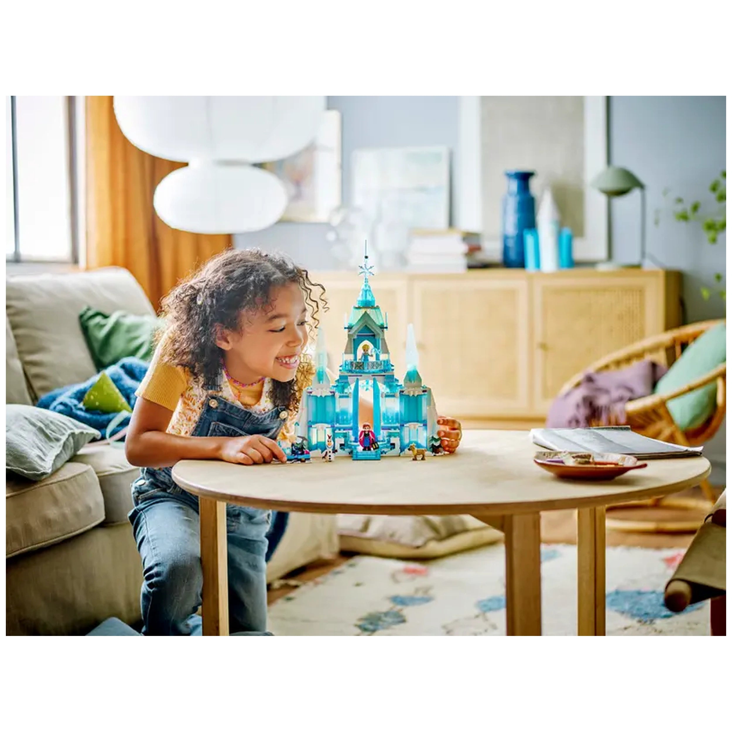 A young girl with curly hair smiles while playing with the LEGO® Disney Frozen Elsa's Ice Palace set on a wooden table. The vibrant blue palace features intricate details and figures from the story, set against a cozy living room backdrop with a couch and decorative elements.