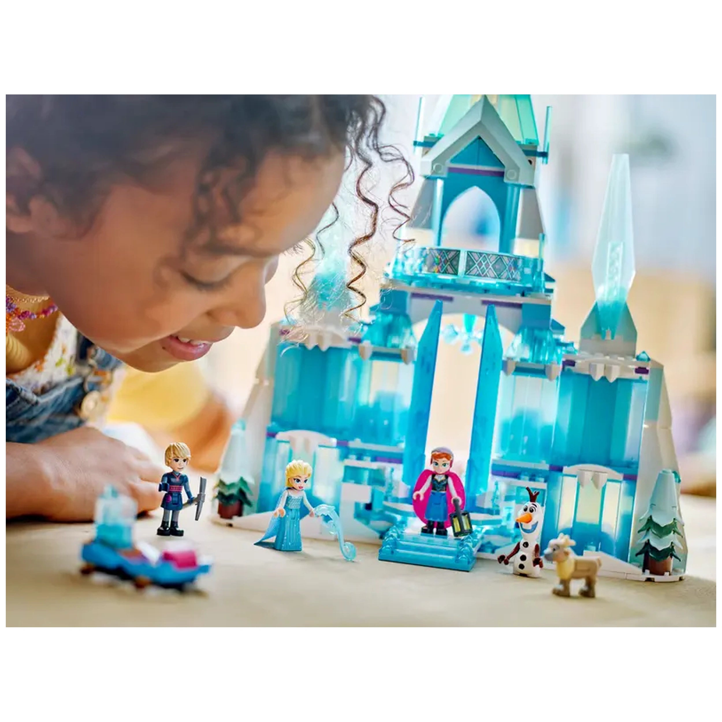 A child leans excitedly over LEGO Disney Frozen Elsa's Ice Palace, featuring a towering icy structure in shades of blue, with Elsa, Anna, Olaf, and a small reindeer figurine nearby. The detailed palace includes whimsical elements like ice formations and a slanted roof, creating a magical play scene.