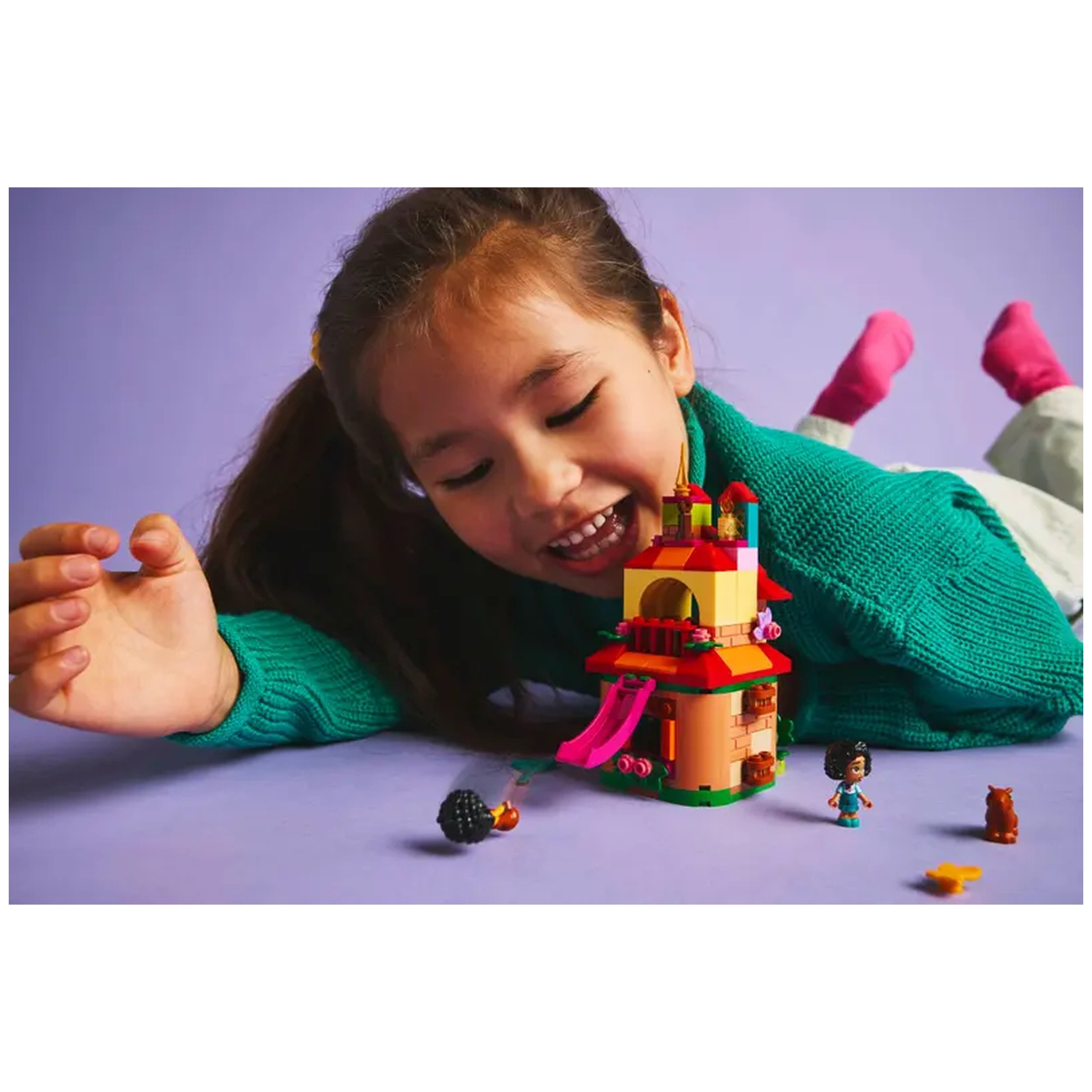 A young girl with a joyful expression plays with a colorful LEGO® Disney Encanto Mini House set on a purple background, surrounded by three character figures and small accessories.