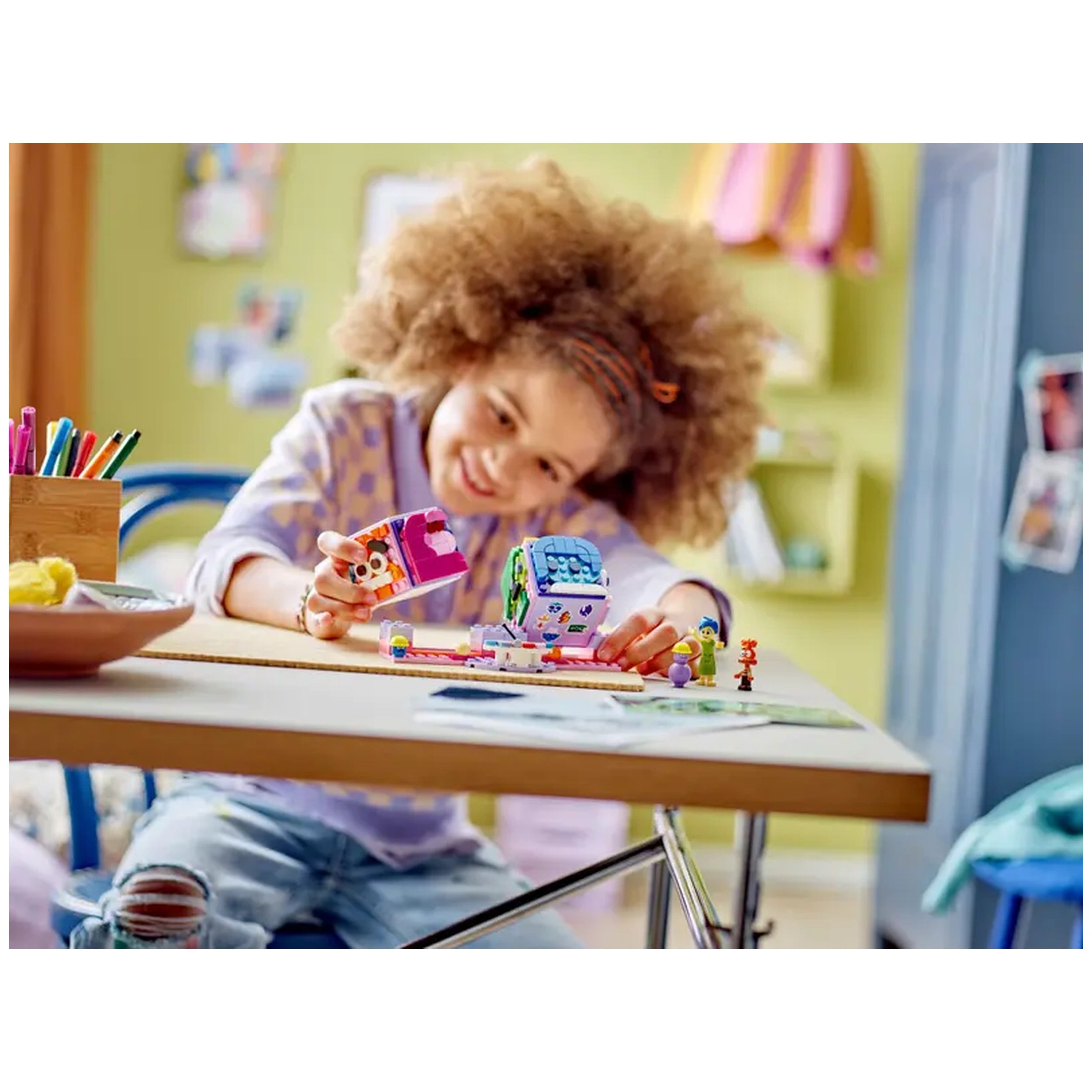A joyful child with curly hair plays with LEGO Disney Inside Out Mood Cubes at a colorful table, holding a mood cube while smiling.