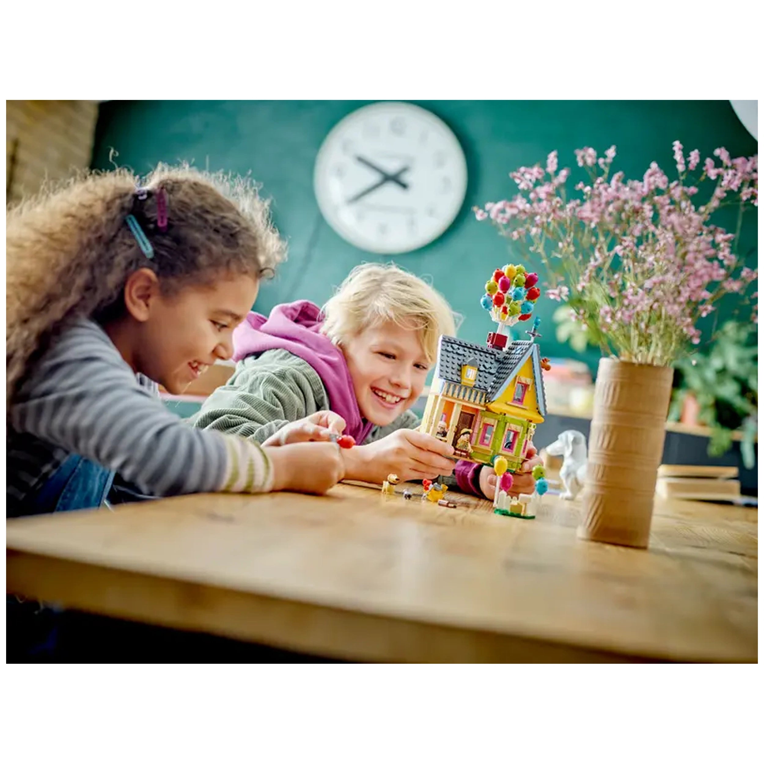 Two children playing with the LEGO® Disney 'Up' House set on a wooden table, smiling as they interact with colorful balloons and minifigures. A vase with flowers sits in the background, adding a vibrant touch to the scene.