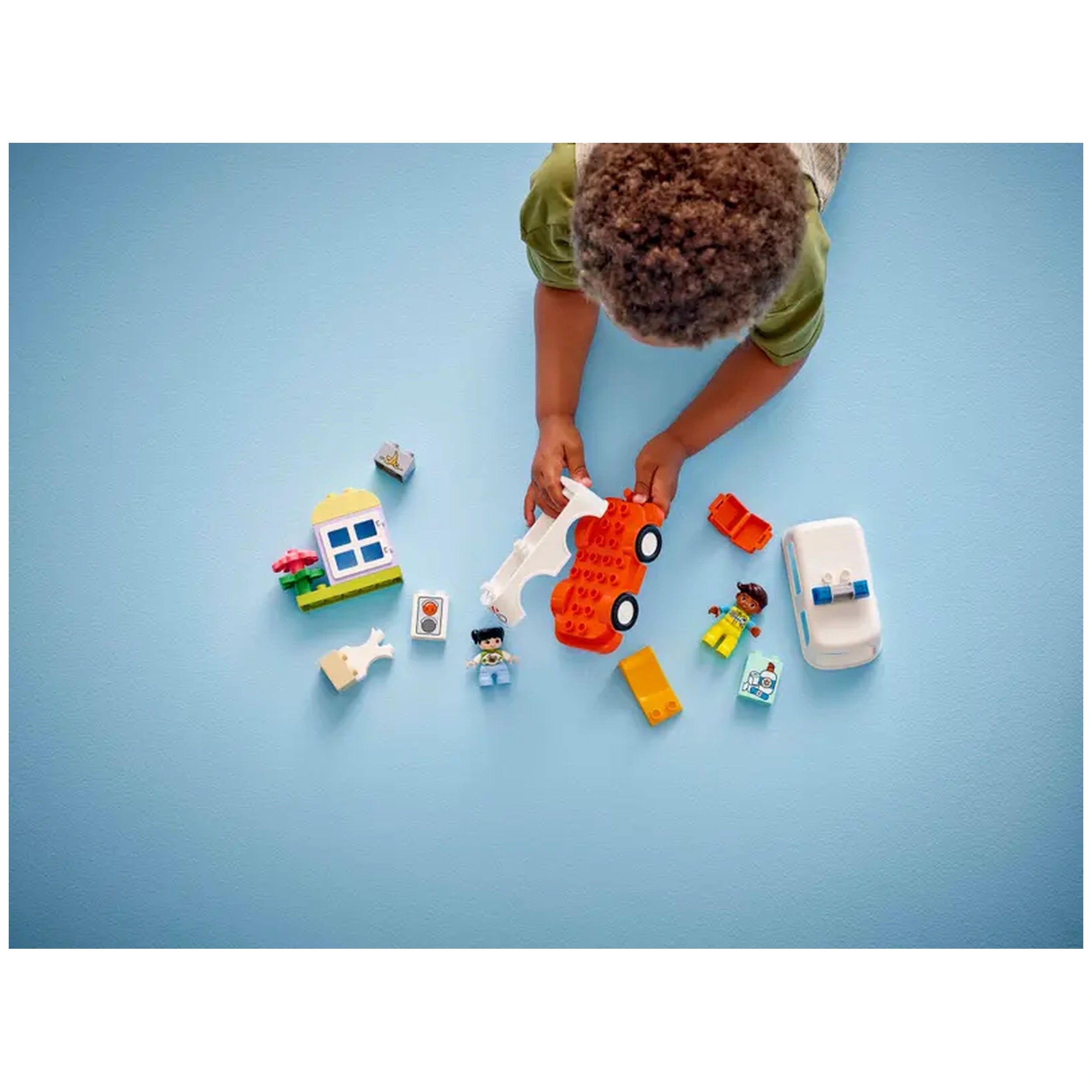 A child plays with the LEGO® DUPLO® Ambulance & Driver set, holding an orange car. Surrounding them are DUPLO figures, a house, and various colorful blocks on a blue background.