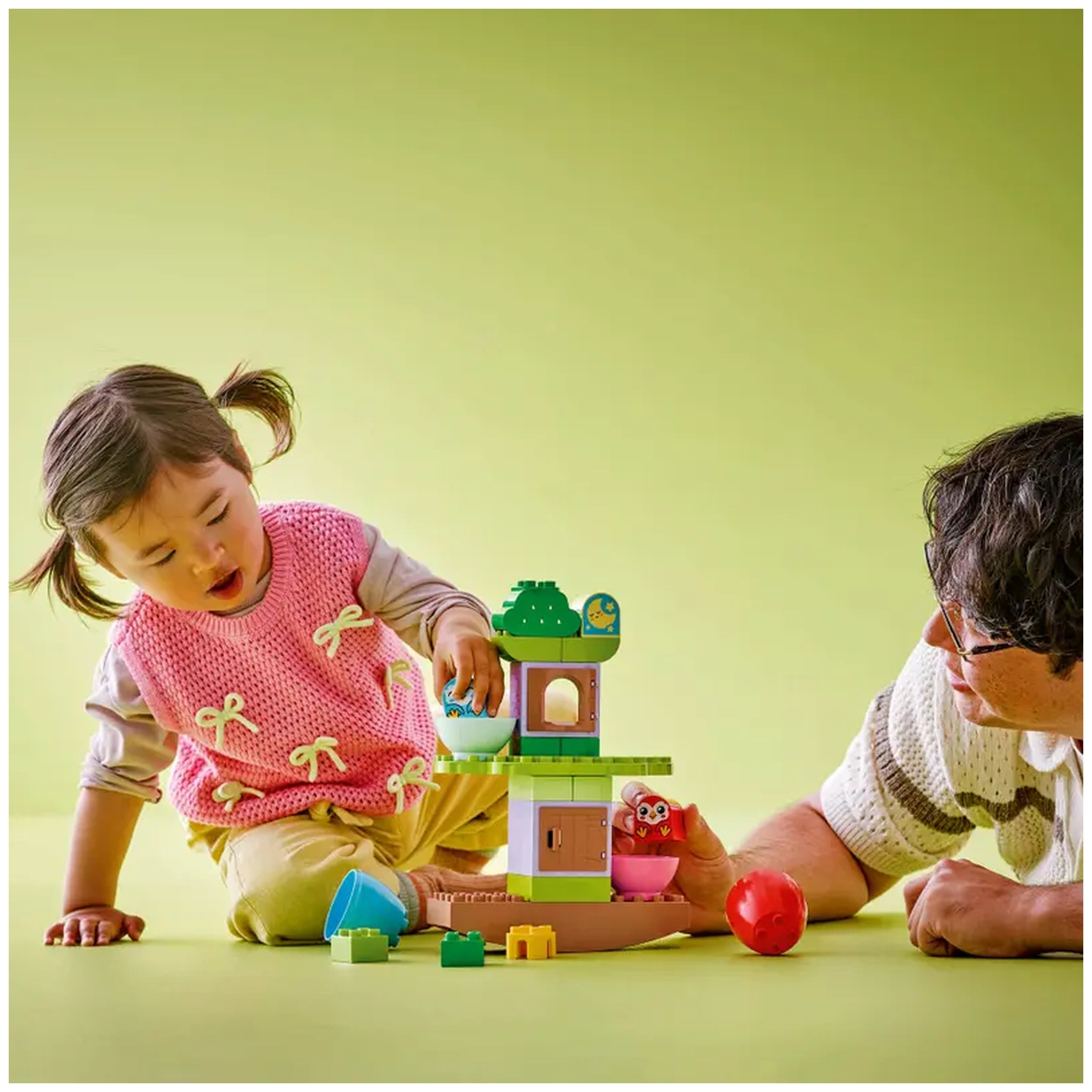A young child in a pink vest plays with the LEGO® DUPLO® Balancing & Stacking Tree set, balancing colorful blocks while a man watches intently.