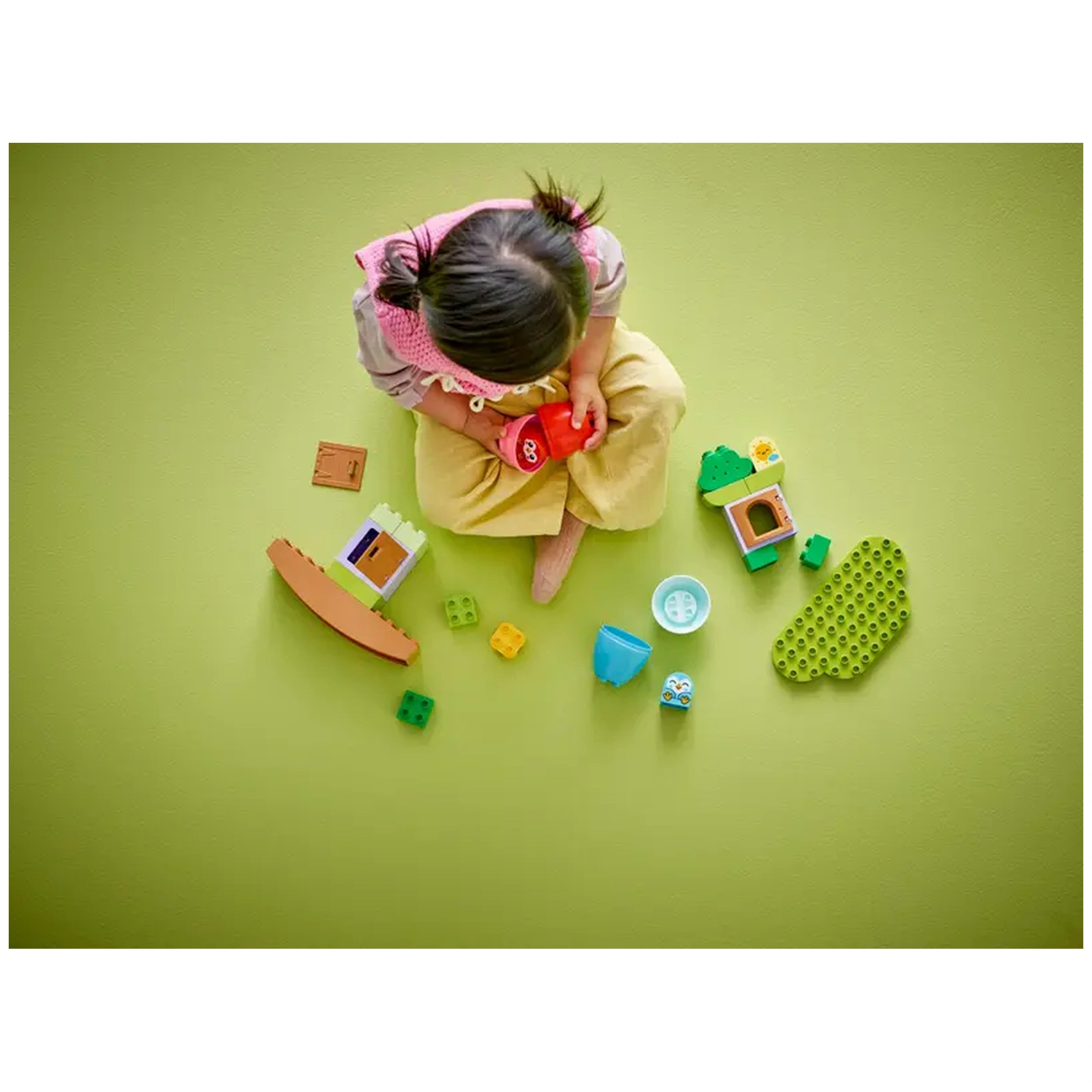 A toddler sits on a green floor, focused on a red toy while surrounded by colorful LEGO® DUPLO® blocks and pieces, including an adorable owl and a tree house.