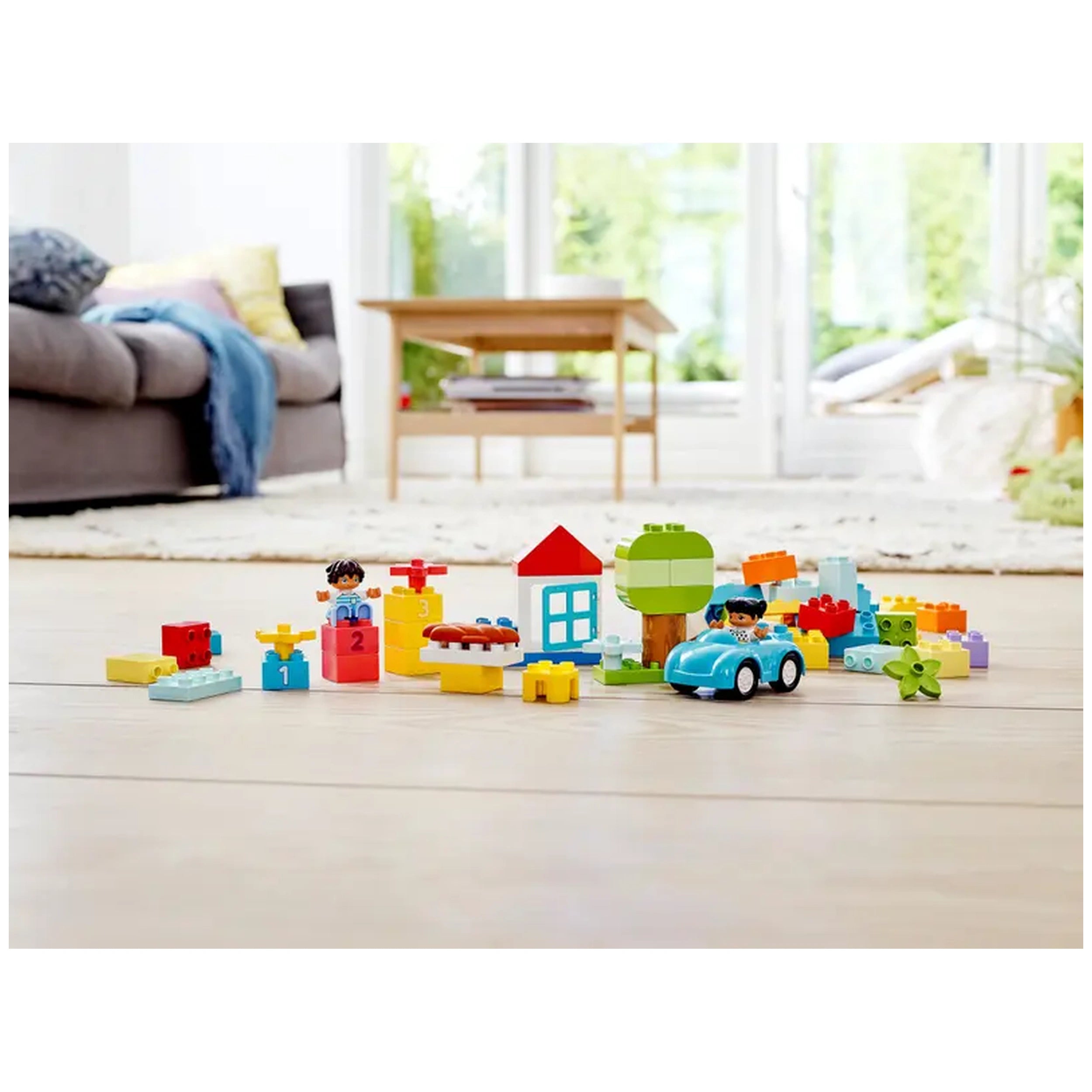 Colorful LEGO® DUPLO® bricks scattered on a wooden floor, featuring a blue car, a house with a red roof, trees, and playful figures, in a bright, sunlit room.