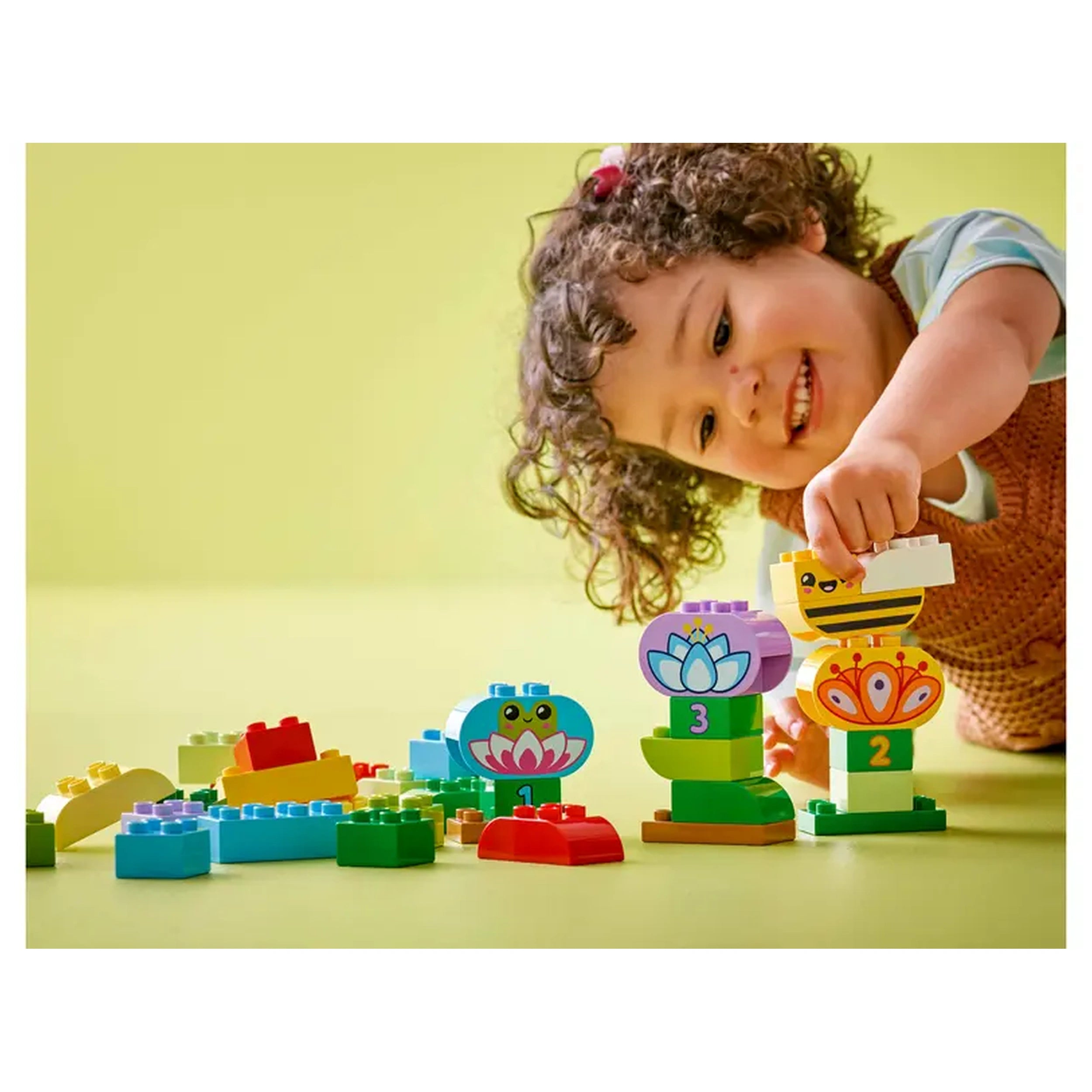 A joyful toddler with curly hair smiles while stacking colorful LEGO® DUPLO® bricks, including flowers and playful animal figures, on a bright green surface.