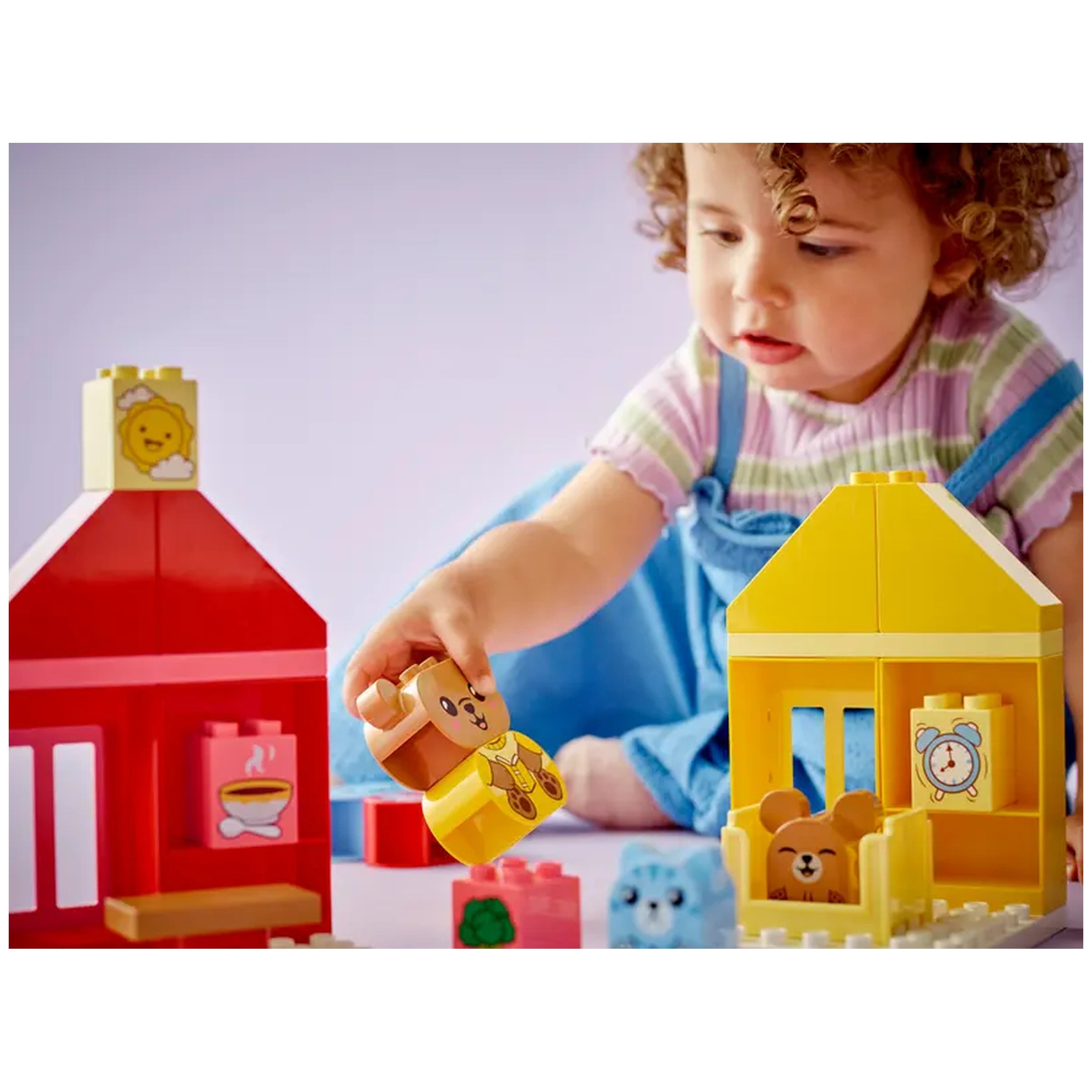 A toddler with curly hair plays with colorful LEGO® DUPLO® blocks, holding a bear figure in one hand. The scene includes a red house and a yellow house with animal figures inside, surrounded by various building pieces on a soft purple surface.