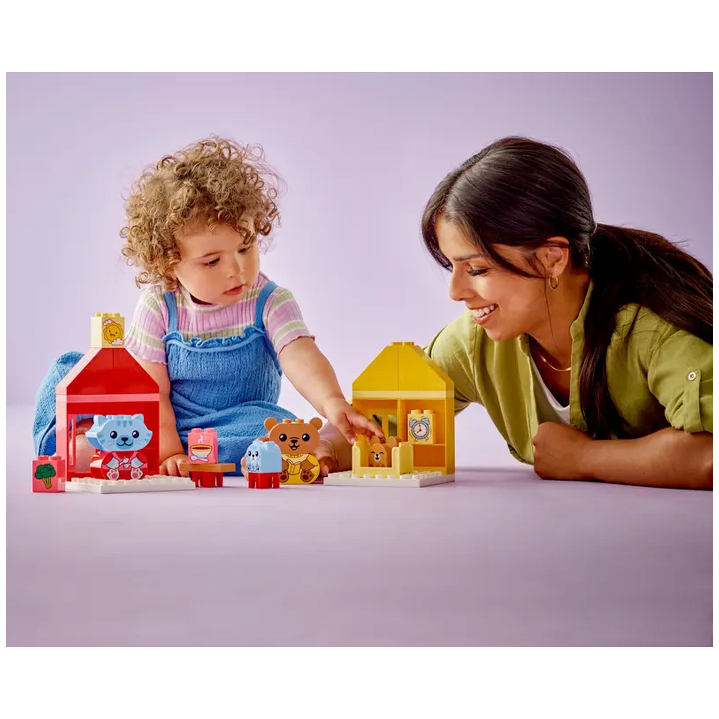 A toddler and a woman engage in play with the LEGO® DUPLO® Daily Routines set. The colorful toys include animal figures and two small houses. Bright pastel background enhances the cheerful atmosphere of interactive play, promoting learning and emotional expression.