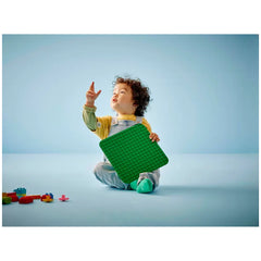 Preschool child sitting on a light blue surface, holding a green LEGO® DUPLO® building plate, looking upwards thoughtfully. Colorful building blocks scattered nearby.