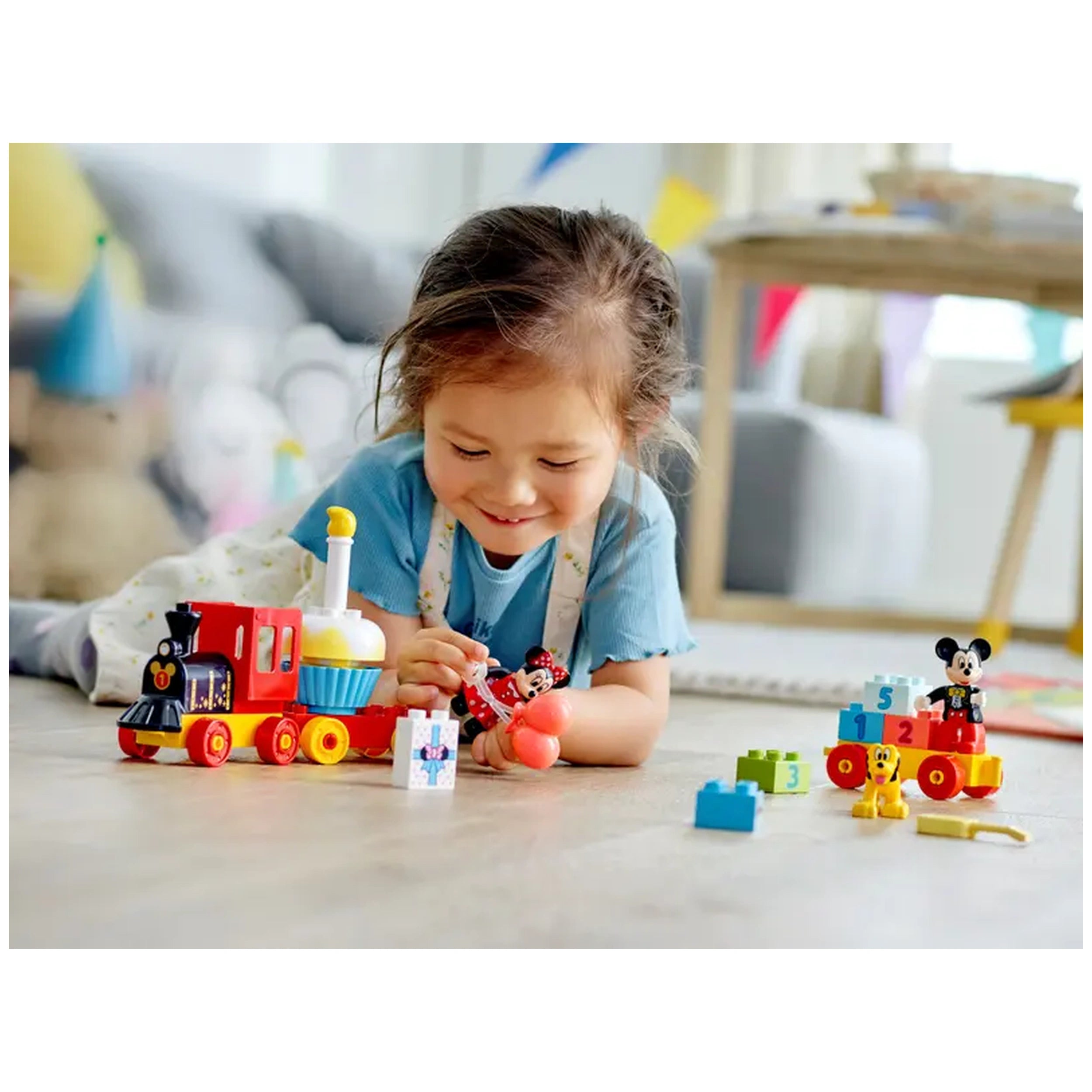 A joyful toddler plays on the floor with the LEGO® DUPLO® Mickey & Minnie Birthday Train set, featuring colorful train cars and cheerful characters. The child, wearing a blue shirt, is engaged with the train while holding a red balloon and a gift box. Bright building blocks are scattered around, enhancing the vibrant scene filled with creativity and fun.