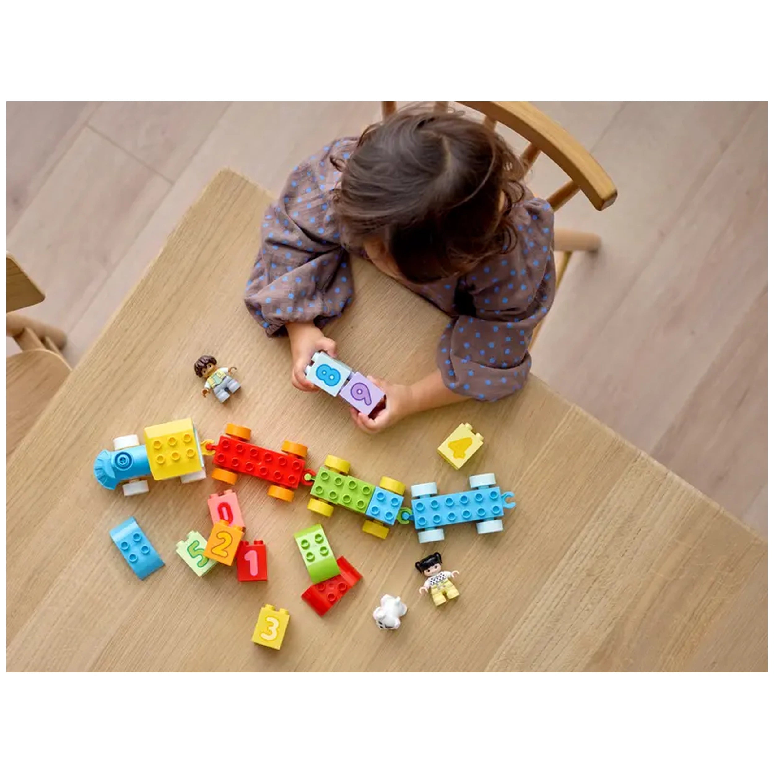 A young child with brown hair, wearing a polka dot shirt, sits at a light wooden table, holding two colorful LEGO bricks showing the numbers eight and a heart. Surrounding them are various LEGO bricks in bright colors and playful figures, including a boy, a girl, and a dog, all part of the LEGO DUPLO Number Train set.