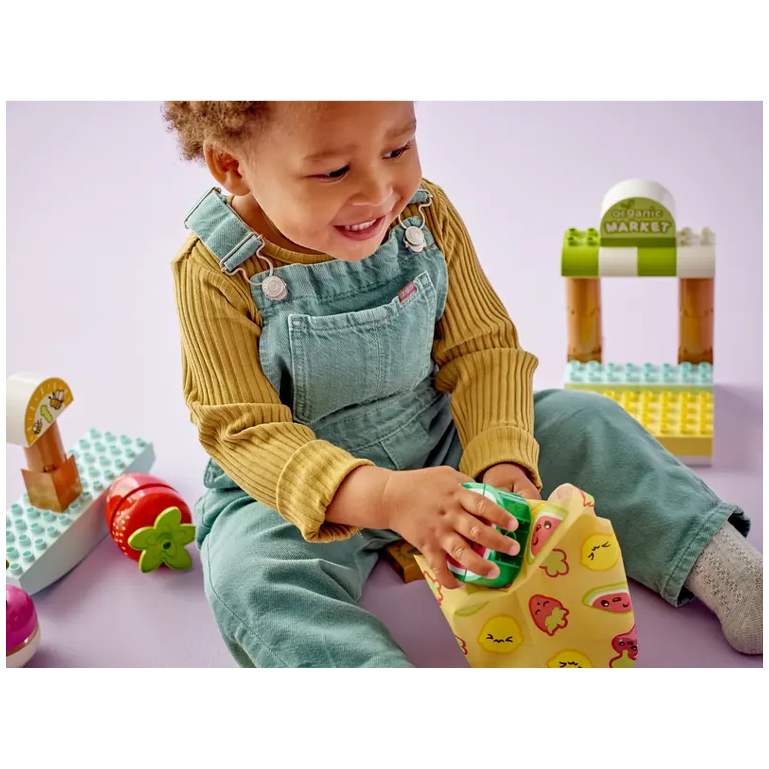 A joyful toddler in a yellow sweater and blue overalls holds a colorful toy while playing beside a LEGO® DUPLO® Organic Market set, featuring vibrant fruits and a market stand, on a soft purple surface.