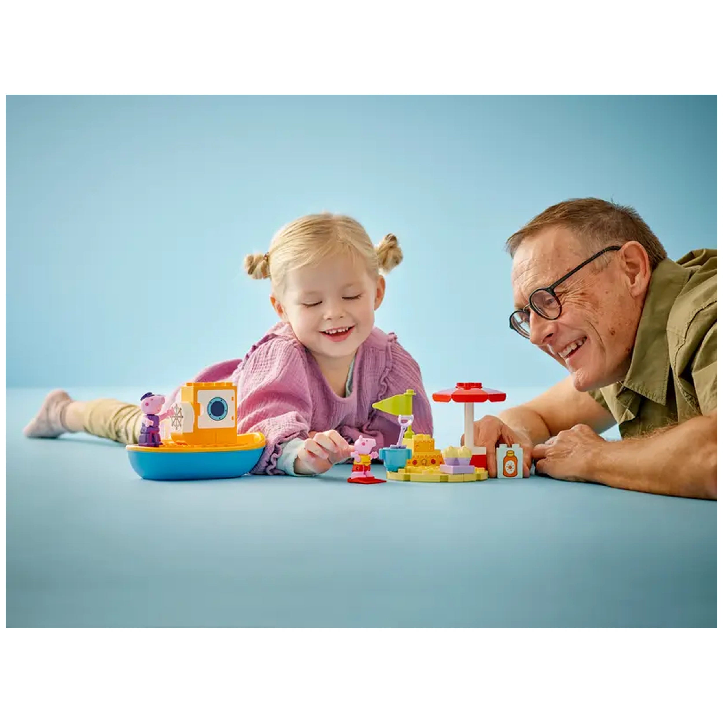 A joyful child and a smiling elderly man play together on a light blue surface, surrounded by LEGO® DUPLO® toys, including a boat, colorful accessories, and characters from Peppa Pig.