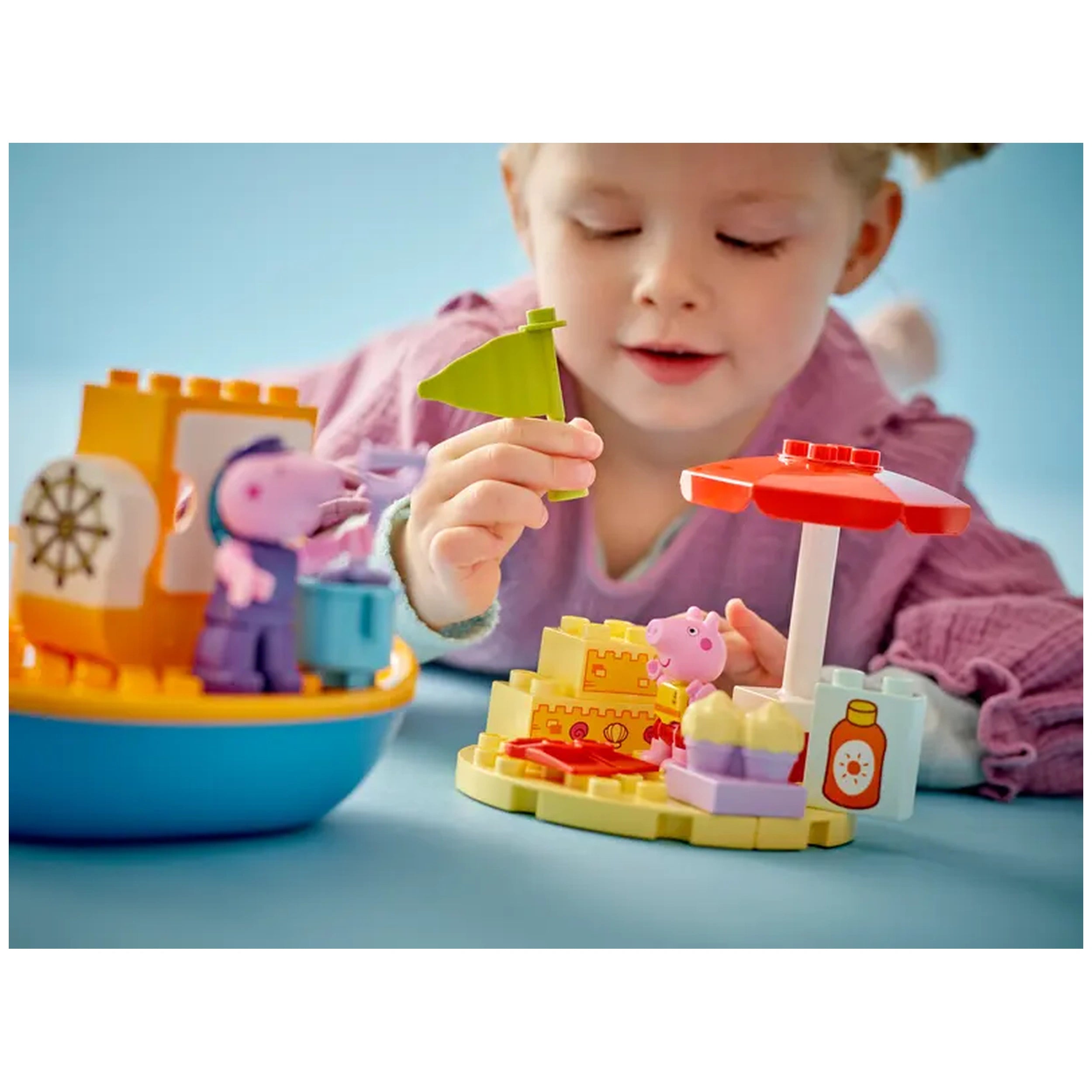 A young child plays with the LEGO® DUPLO® Peppa Pig Boat Trip set, holding a green flag. The colorful toy features a boat, a beach scene with a red and white umbrella, and figures of Peppa and Grandpa Pig among sandcastles and accessories.