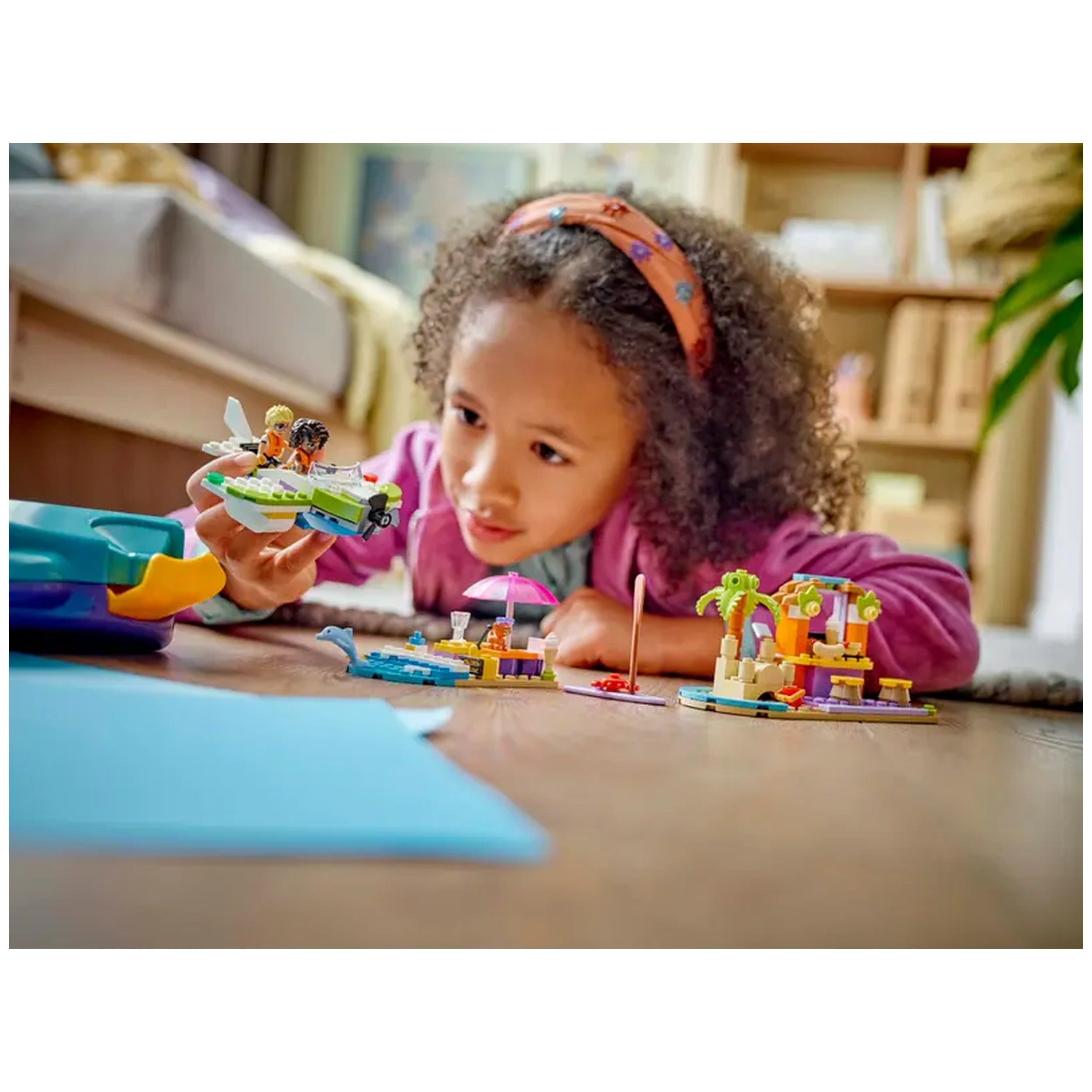 A child with curly hair examines a colorful LEGO® Friends seaplane while surrounded by beach-themed building sets, including a café and beach cabana.