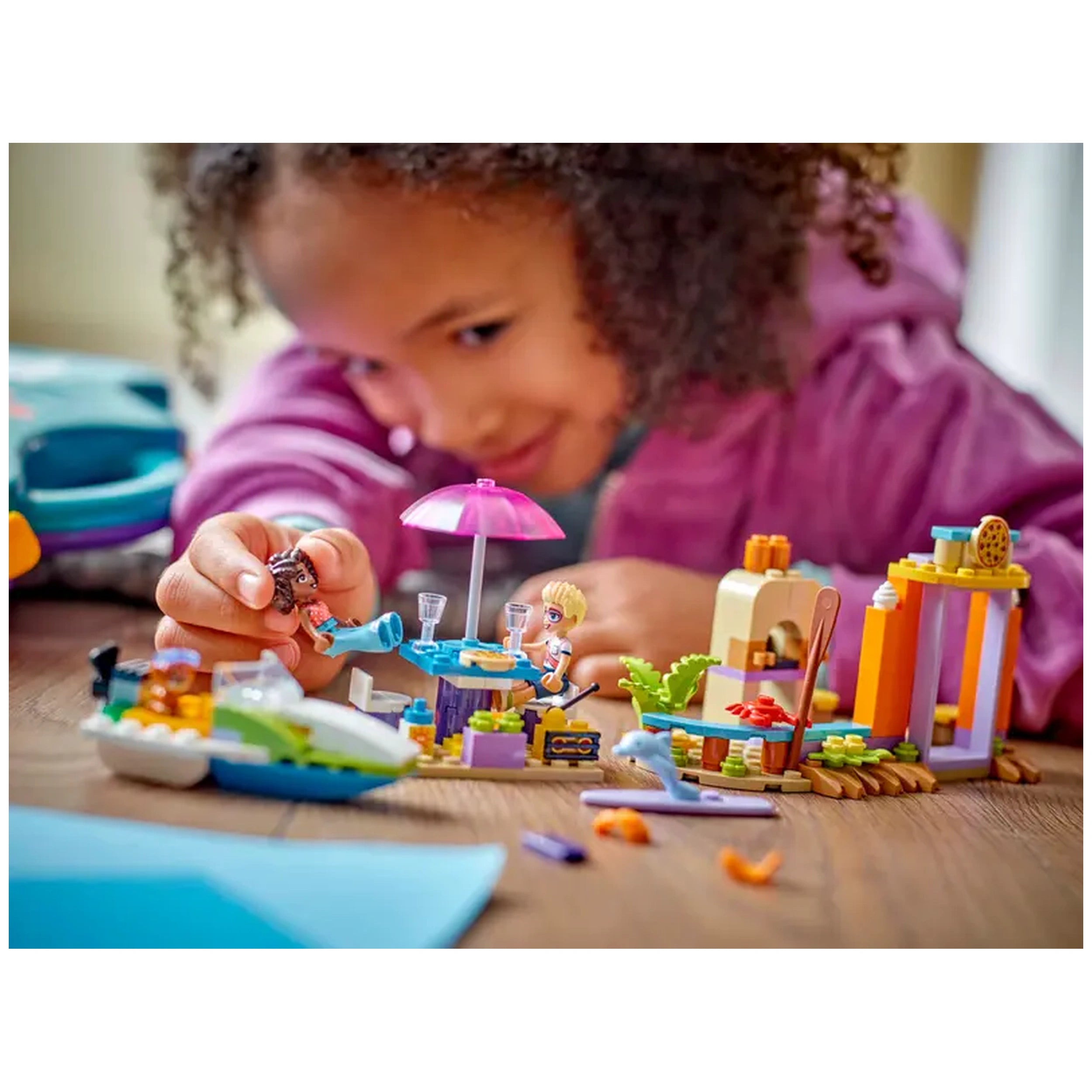 A young child plays with colorful LEGO® Friends toys, constructing a beach scene with a pink umbrella table, mini figures, and a small boat.
