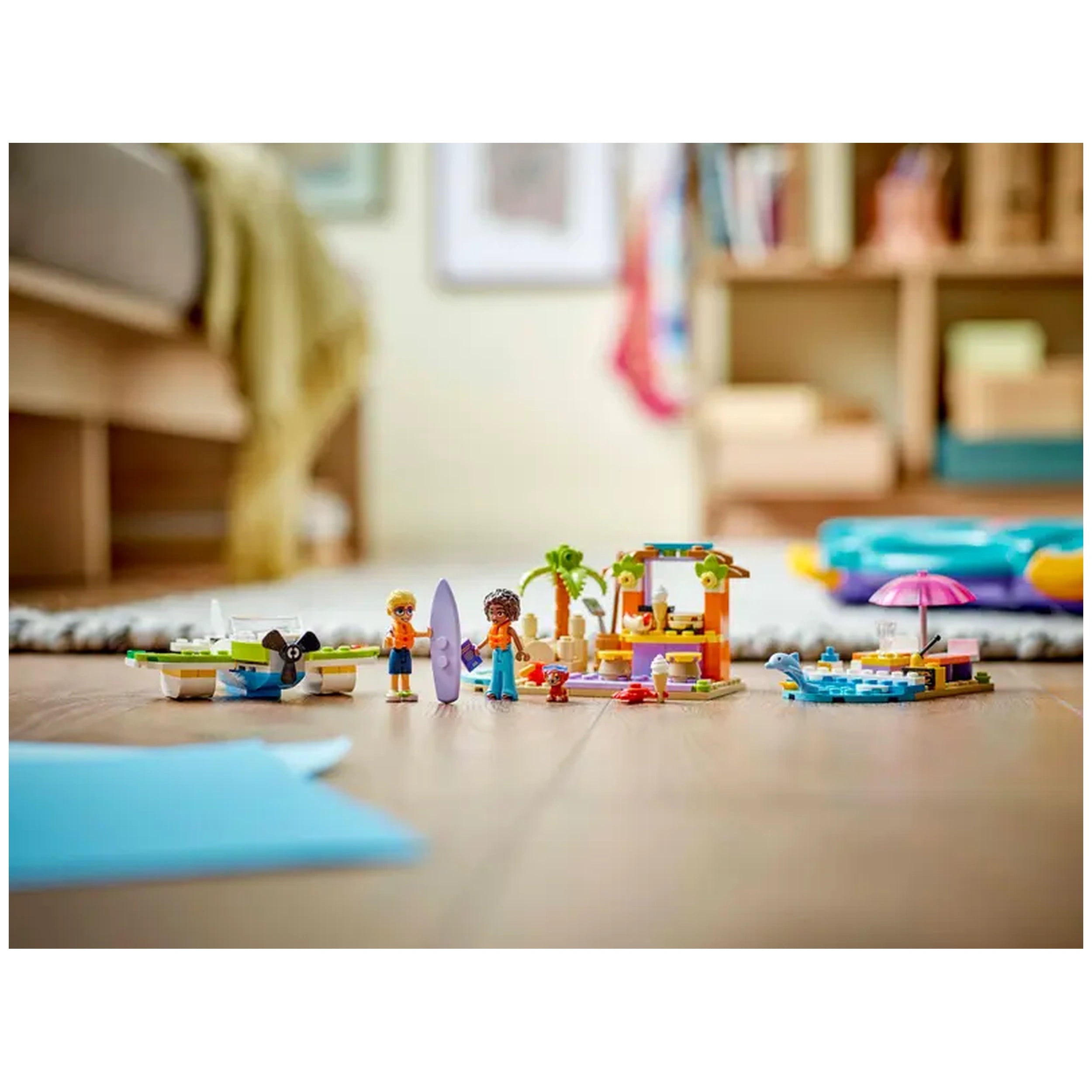 Colorful LEGO® Friends beach scene with two figures holding a surfboard, a café, a seaplane, and a small boat under a pink umbrella.