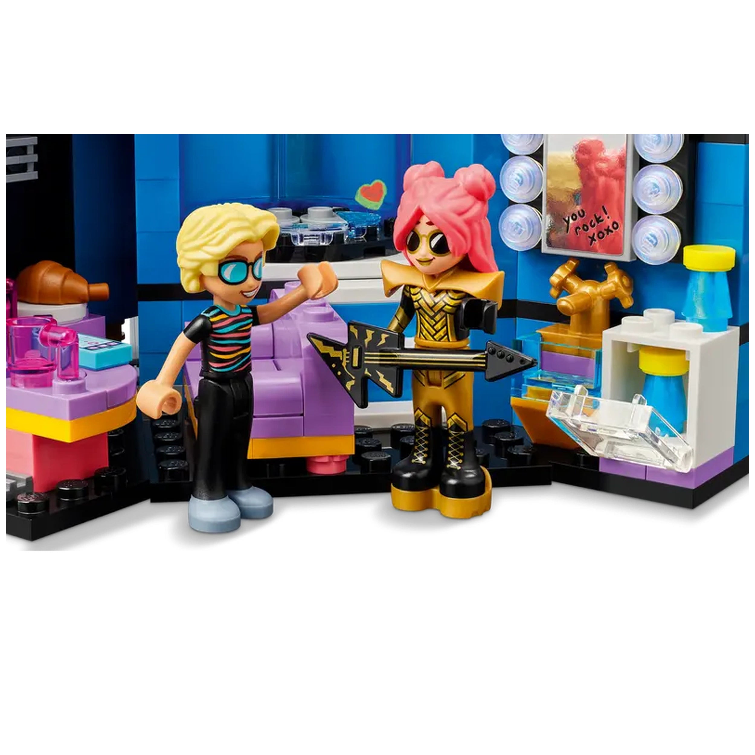 Two colorful mini-dolls in a music-themed playset, one holding a guitar and the other gesturing excitedly. Bright pink and gold outfits contrast with a purple stage backdrop featuring musical accessories.