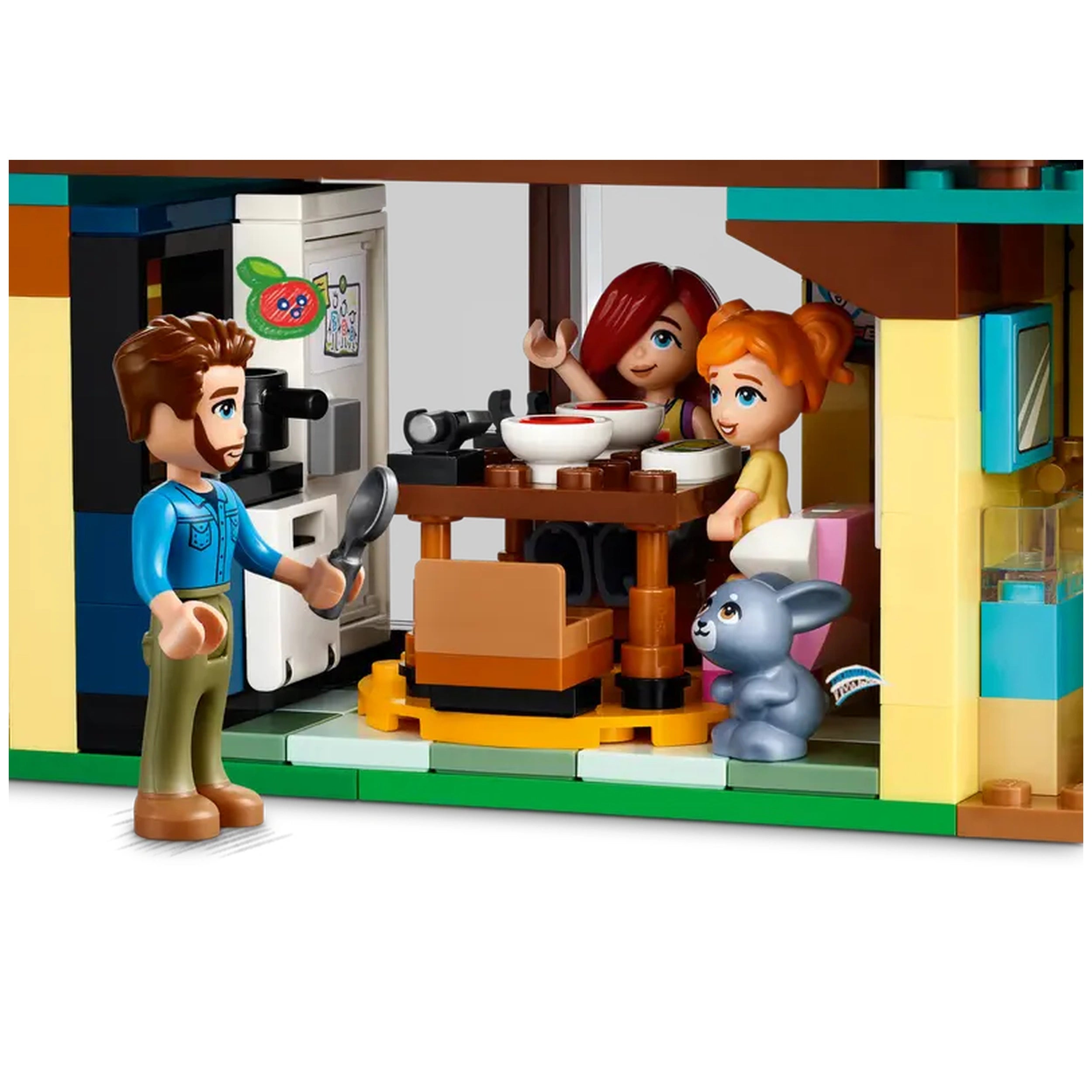 A LEGO scene featuring a friendly male figure holding a spoon, engaging with two girls at a dining table with bowls. A small blue pet rabbit sits nearby, and a refrigerator is visible in the background.
