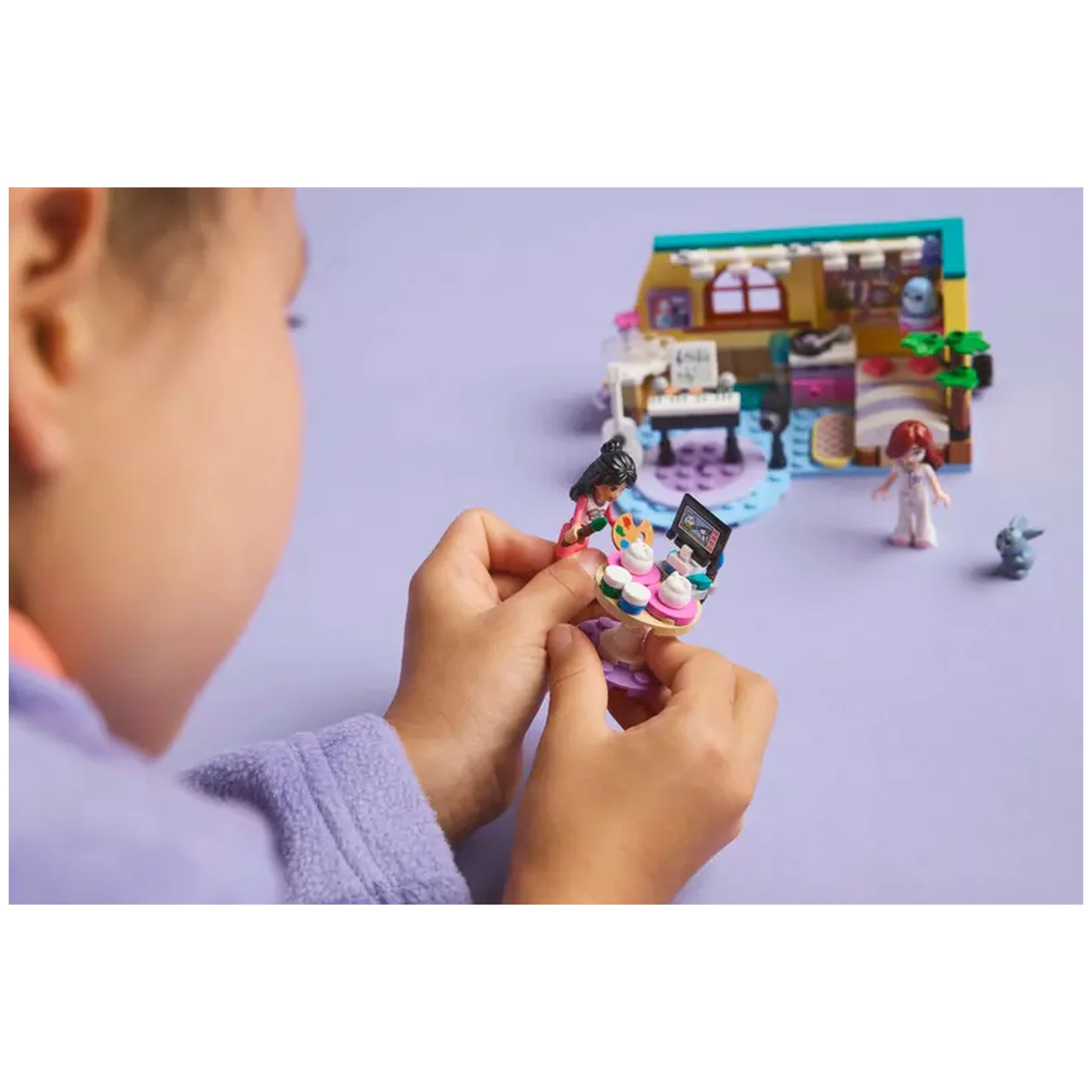 A child in a cozy purple robe holds a LEGO Friends minidoll with a colorful cupcake display, in front of Paisley’s Room set.