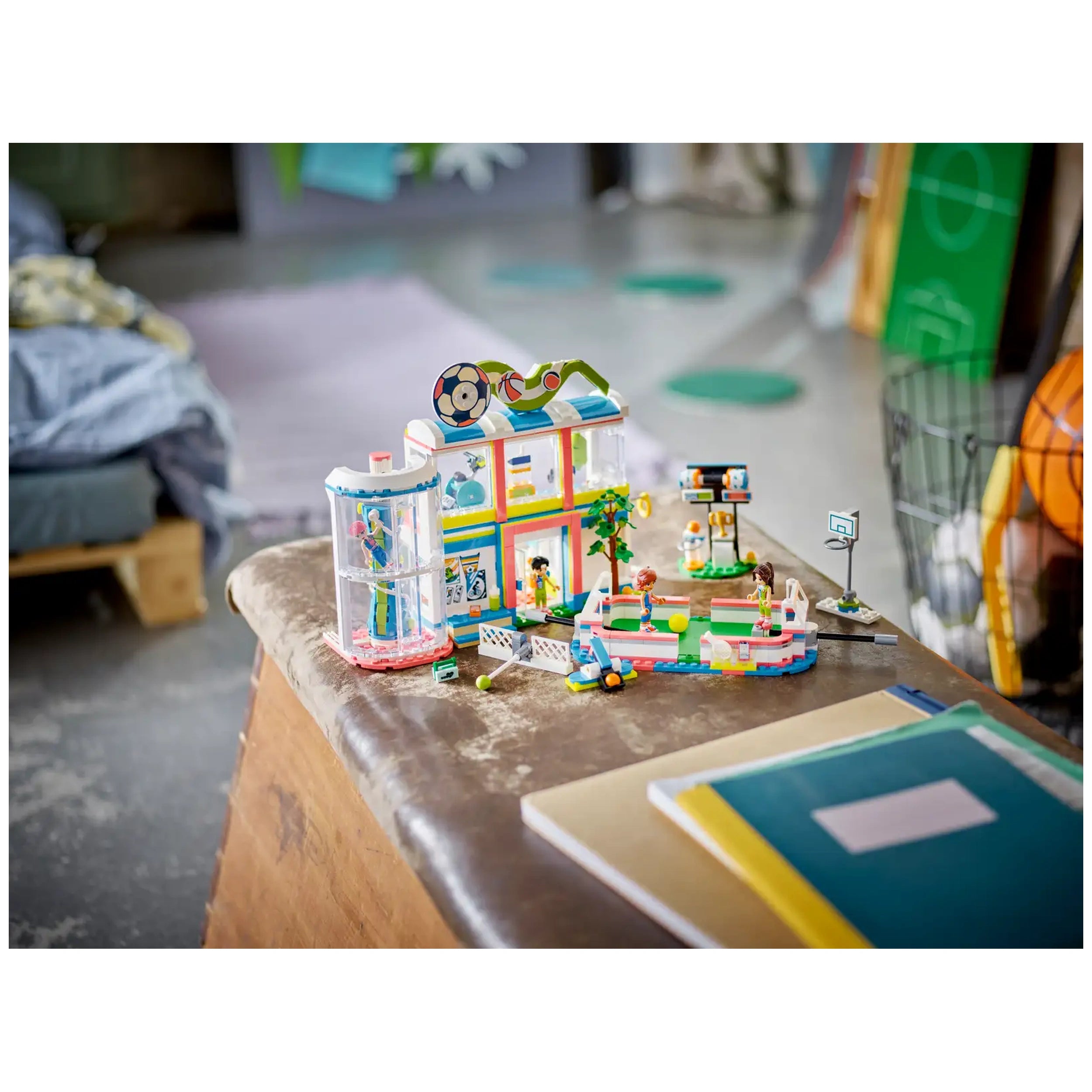 Colorful LEGO® Friends Sports Center set on a wooden table, featuring a variety of sports scenes with mini-dolls playing soccer and basketball, surrounded by accessories like balls and nets.