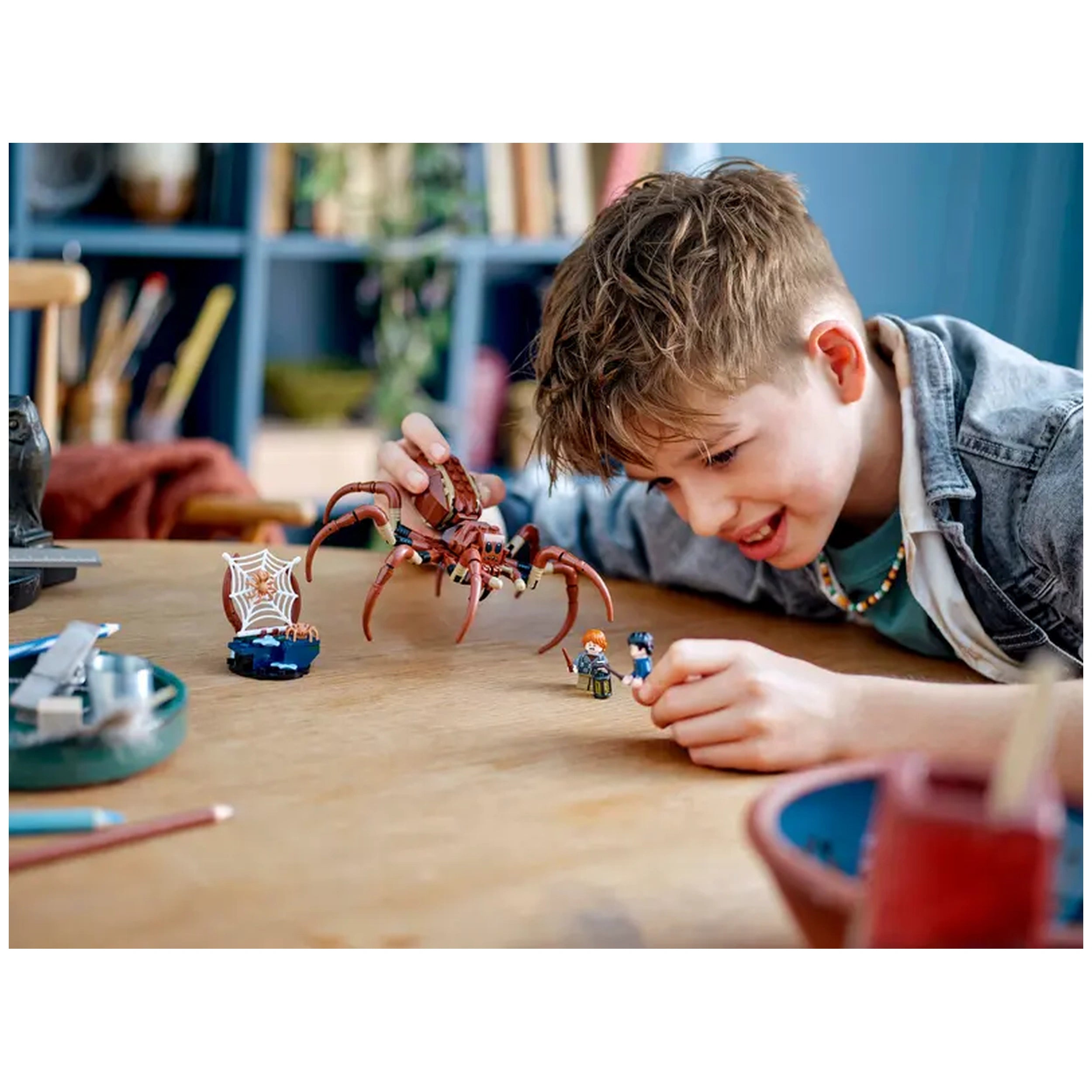 A smiling boy with short hair plays on a wooden table, holding a large LEGO Aragog spider figure. Two LEGO minifigures of Harry Potter and Ron Weasley are in front of him, along with a small spider web accessory. Colorful pencils and a round bowl are nearby, creating a playful and creative atmosphere.