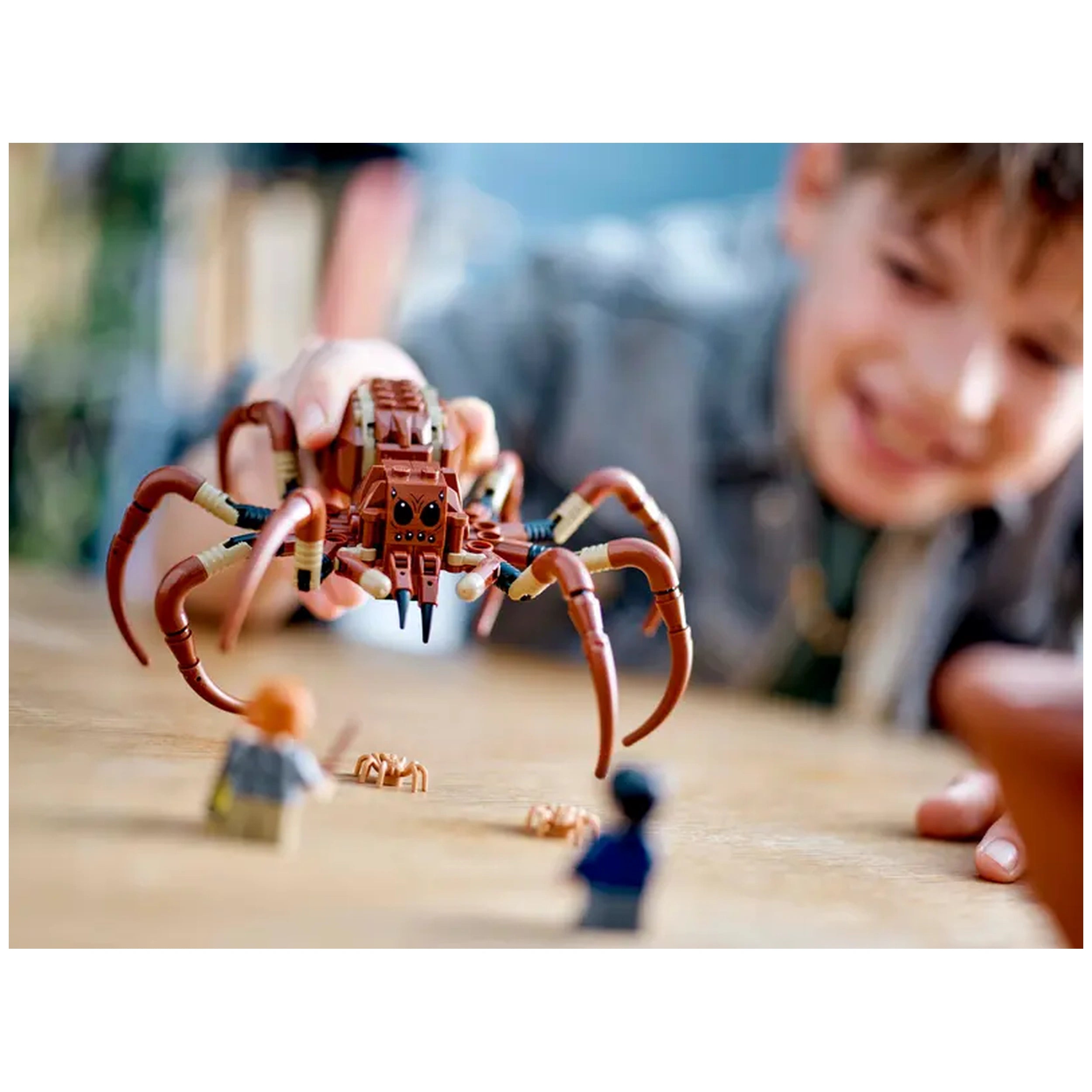 A child excitedly interacts with a large, brown LEGO Aragog spider figure, poised above smaller minifigures of Harry Potter and Ron Weasley on a wooden table.
