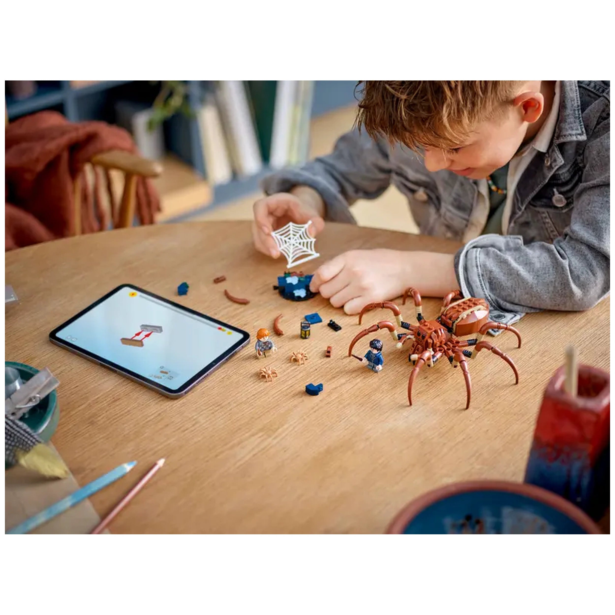 A child builds the LEGO Harry Potter Aragog set on a wooden table, assembling a spider figure and minifigures with an interactive tablet nearby.