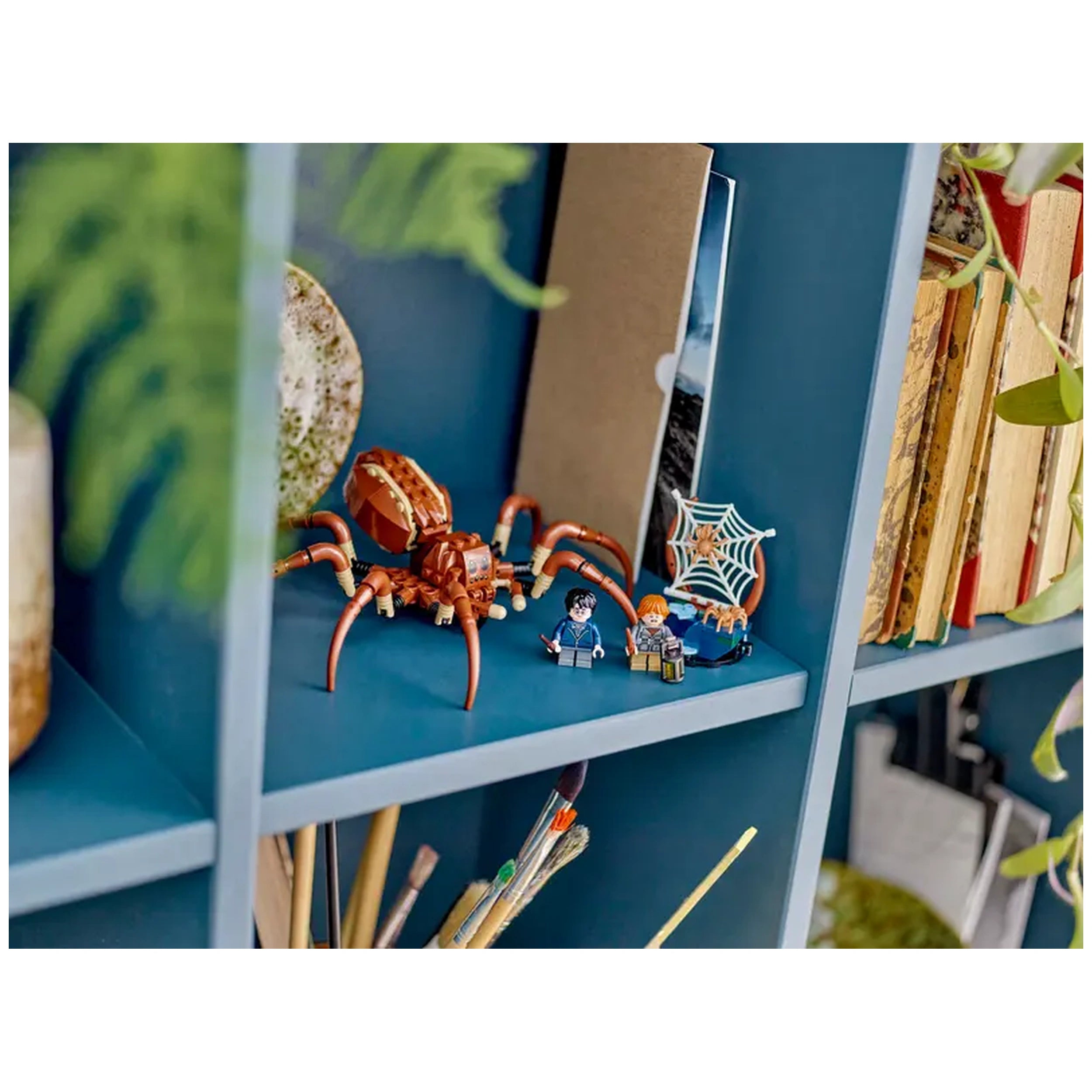 LEGO® Harry Potter™ Aragog figure in brown, with posable legs, positioned beside minifigures of Harry Potter and Ron Weasley, on a blue shelf adorned with books and a decorative plant. A spider web accessory is also displayed.