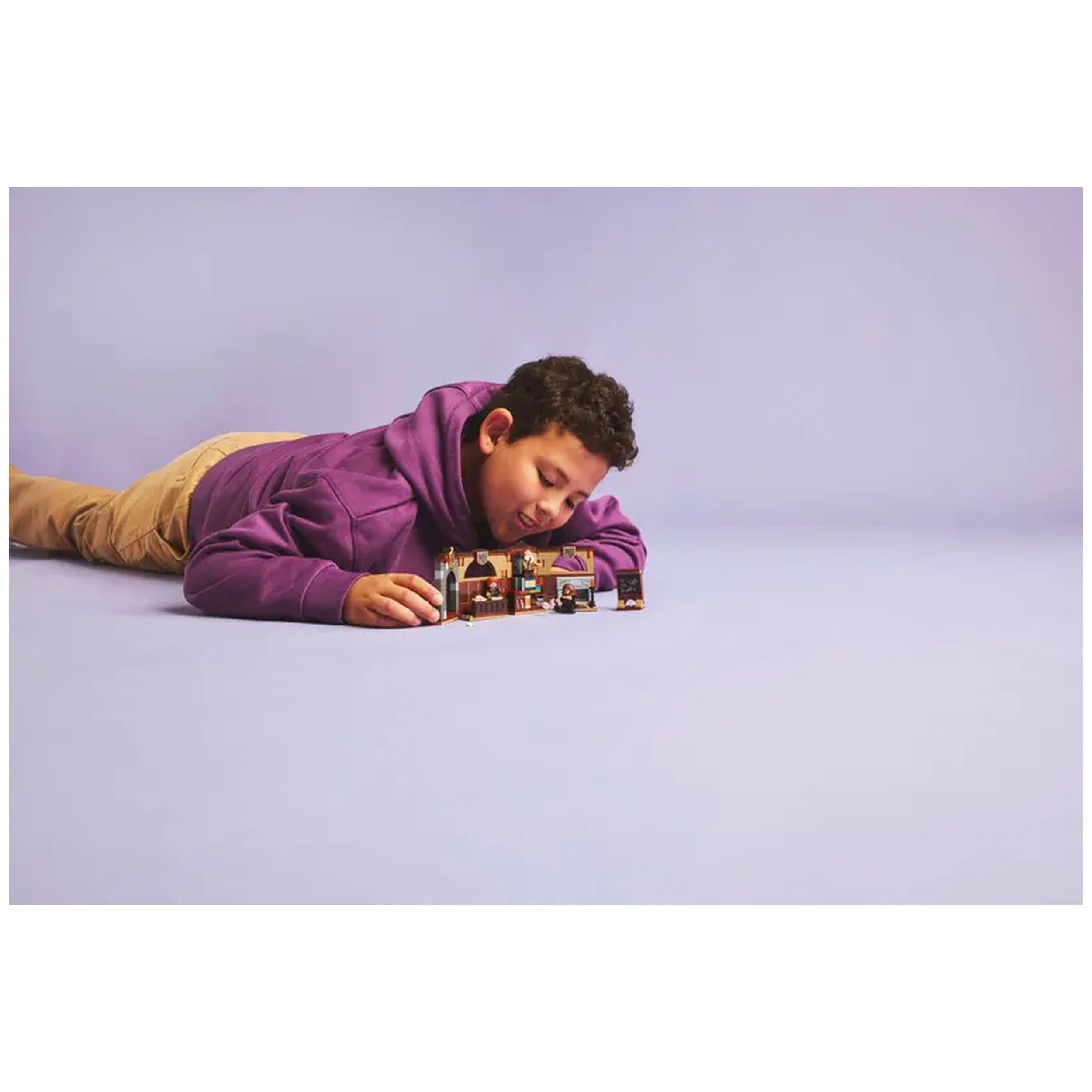 A child in a purple hoodie lies on a light purple surface, eagerly engaging with a LEGO Hogwarts Castle: Charms Class playset, featuring intricate classroom details.